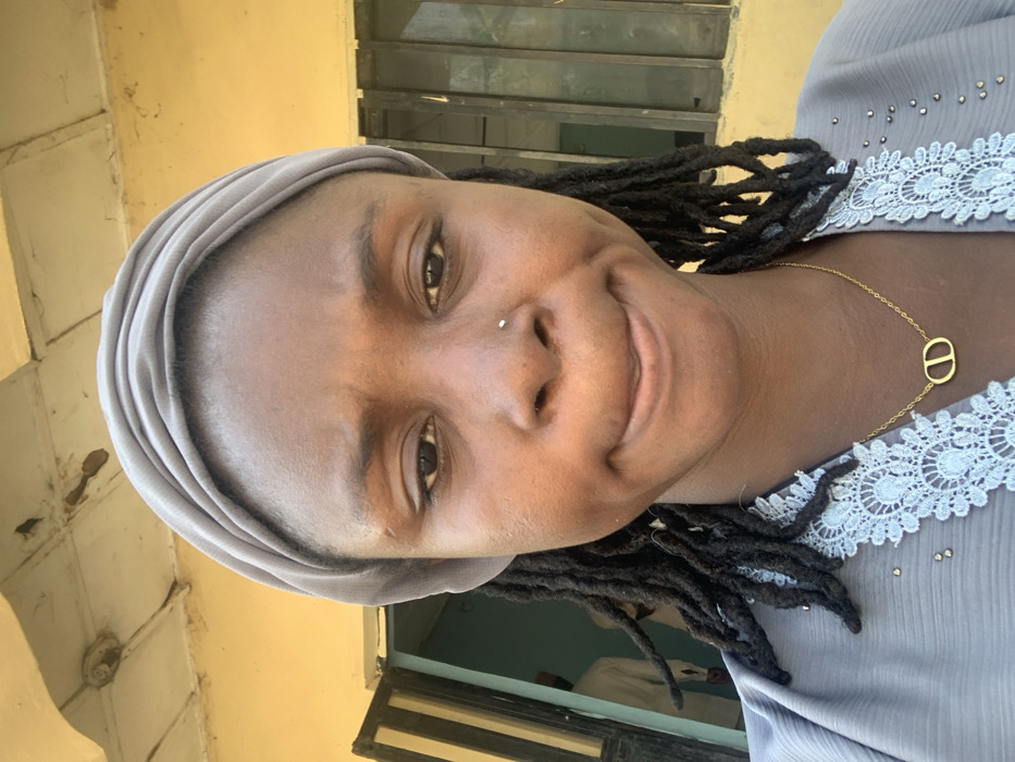 The image captures a close-up portrait of a young adult female in the city of Bojude, Nigeria. The subject, of dark complexion, wears a light grey fabric headwrap and has dark dreadlocks partially visible. Her expression is characterized by a furrowed brow and pursed lips, eyes directed at the camera. A small, light-colored stud is present on her right nostril. She wears a light grey garment with light blue lace trim and a thin gold-colored necklace featuring an interlocking 'CD' shaped pendant. The background reveals a yellow-painted building exterior. To the left, an open doorway displays a light blue interior and a partially visible, blurred male figure in light-colored attire. To the right, a window with dark framing is visible. An overhead ceiling structure, also light-colored, is partially within the frame above the subject.