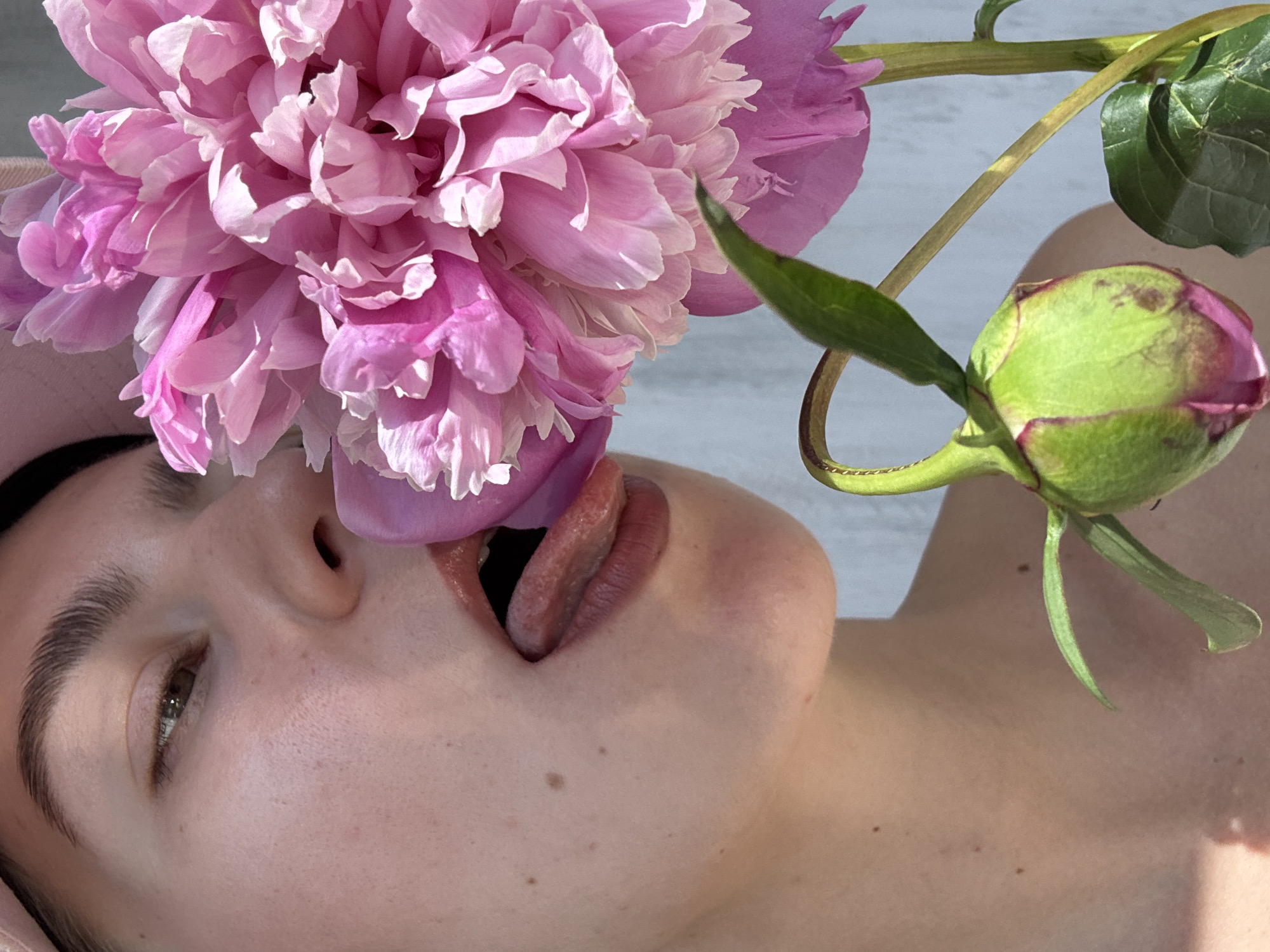 A close-up, profile view of a person's face and upper body is captured, positioned to the left of a large, vibrant pink peony flower. The person's mouth is slightly open, with their tongue extended and touching the petals of the flower. Their eyes are partially closed, and they are wearing a pink cap. The skin exhibits freckles and subtle pores. The peony is in full bloom, with delicate, layered petals, and a smaller, green bud is visible below it. The background is a textured, light blue wall, with sunlight casting shadows on the person's skin. The scene is devoid of any identifiable urban landmarks or specific environmental indicators that would suggest a city location.