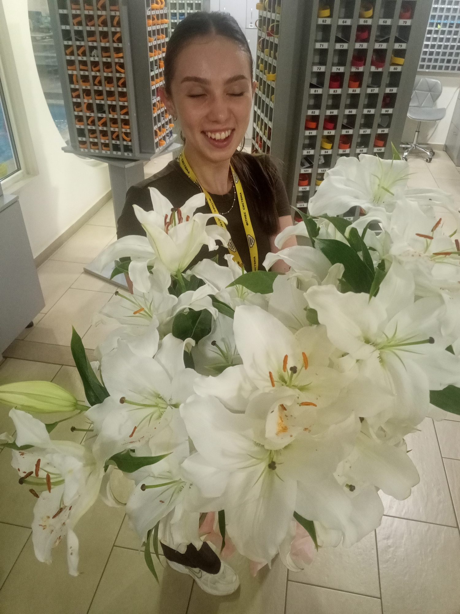 This media file shows a young woman holding a large bouquet of white lilies. She is smiling broadly with her eyes closed, conveying happiness or joy. She is wearing a dark t-shirt with a yellow lanyard around her neck that has text on it. Behind her, there is a wall with many numbered cubbies or shelves, some containing colored items. To her left, there is a window and part of a grey cabinet. To her right, there is a grey office chair. The floor is tiled. The scene appears to be indoors, possibly in a locker room, storage area, or a place associated with fitness activities, given the cubbies and the text on the lanyard. The lighting suggests it could be daytime. The text on the lanyard is partially visible and appears to be in a Cyrillic alphabet, with "ФІТНЕС СИСТЕМА" being the most legible part. The cubbies are numbered from 60 up to 111, and some contain colorful items like red, yellow, and black.