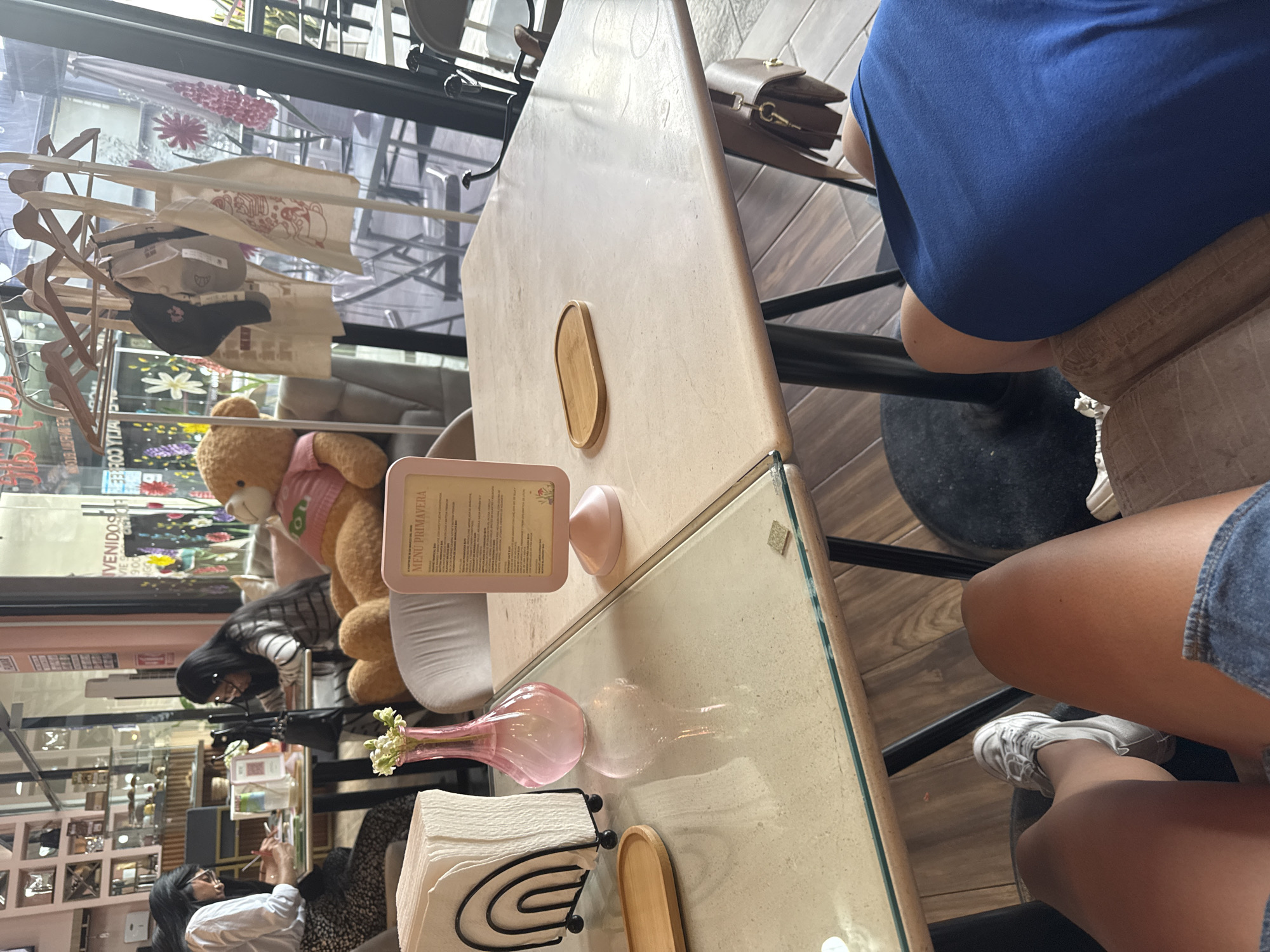 The image shows a table in a cafe in Puebla, Mexico. A teddy bear wearing a pink shirt is seated at the table next to a menu. Behind the bear, a woman wearing a striped shirt and glasses is looking down at something on the table. Another woman wearing a white shirt and black patterned skirt is seated at the table, possibly writing. A vase of flowers, a stack of napkins, and other small objects are on the table. A person wearing a blue shirt is seated at the table.