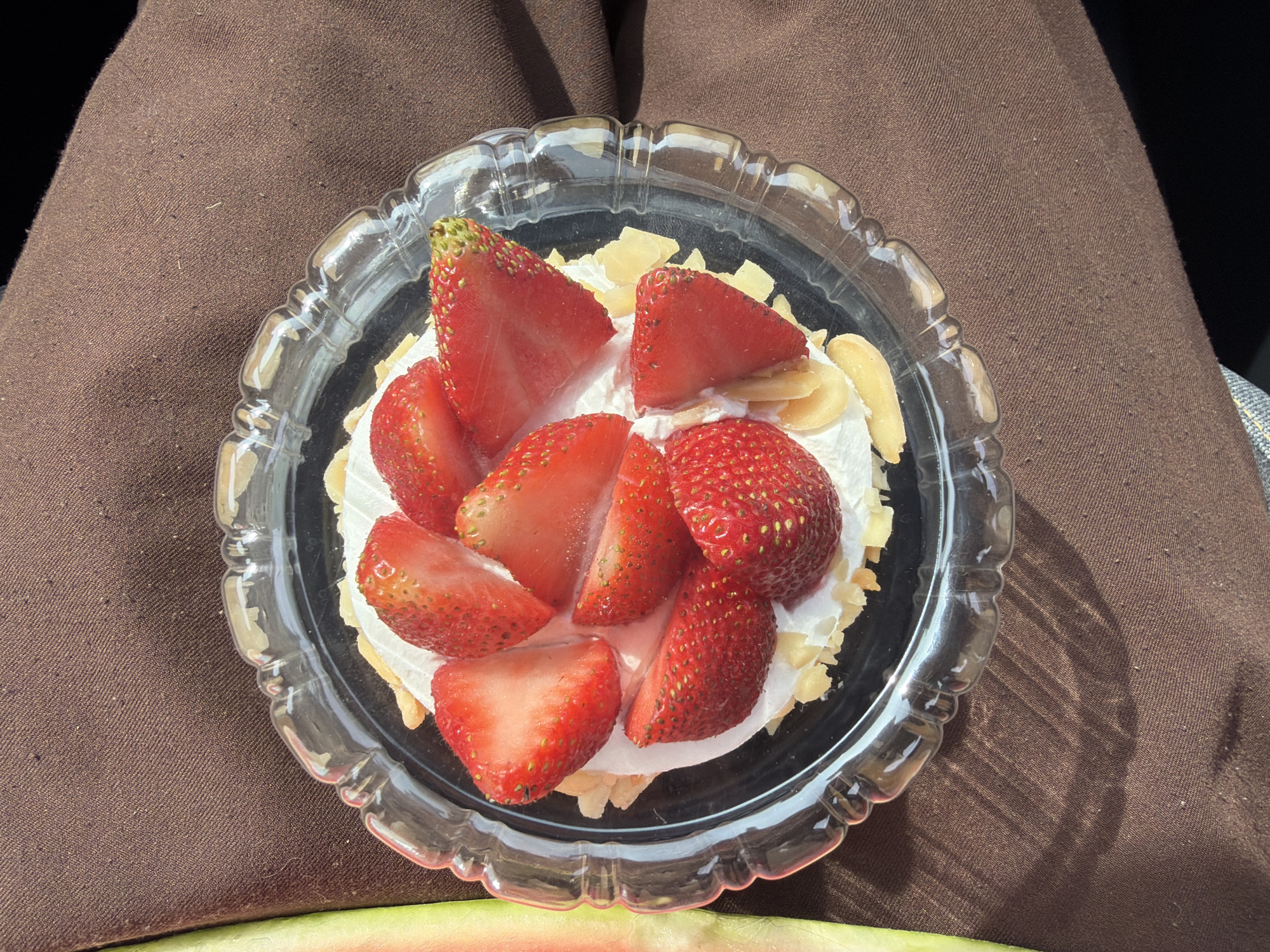 A high-angle, close-up shot shows a clear glass dessert bowl filled with a strawberry shortcake. The cake is topped with whipped cream, sliced almonds, and several sliced strawberries. The dessert bowl sits on a brown surface, possibly clothing, with a hint of green from what appears to be a watermelon visible at the bottom. The photo was taken during daylight hours. The location is not specified.