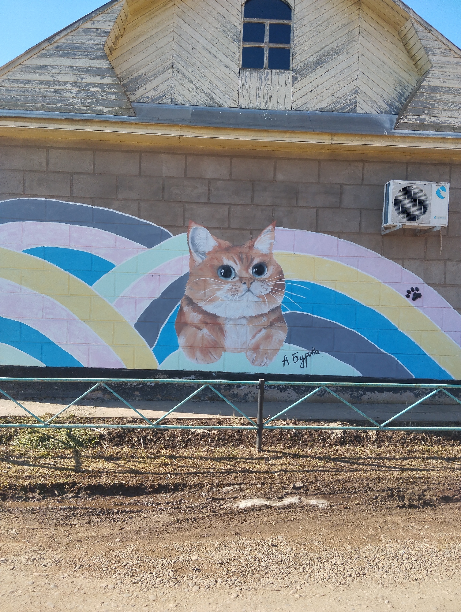 An outdoor, eye-level shot captures a vibrant cat mural painted on the concrete block wall of a weathered building, likely in Krasnogorskoye, Russia. The building features an upper section with light-colored, peeling wooden siding arranged in a chevron pattern on its gable, and a small, arched window with dark panes. Below the gable, the wall is constructed from grey cinder blocks.
The prominent mural depicts a large, fluffy orange cat with striking blue eyes, peeking from behind a series of undulating, colorful stripes. These stripes, resembling hills or a stylized rainbow, are painted in pastel shades of pink, yellow, blue, and grey. A small black paw print is visible on the upper right side of the mural. Below the cat's head, the artist's signature, "А. Бурова" (A. Burova), is written in cursive. To the right of the mural, a white air conditioning unit with a blue logo is mounted on the concrete wall.
In the foreground, a low, green metal fence with a diamond pattern spans the width of the image. Beyond the fence, the ground is a mix of muddy dirt, sparse dry grass, and small rocks, with some visible tire tracks or ruts. The very bottom of the frame shows a wider, gravelly dirt path or road. The scene is brightly lit by natural sunlight from a clear blue sky, casting distinct shadows from the fence onto the ground, suggesting it is daytime. The overall impression combines the utilitarian architecture with a whimsical artistic touch.