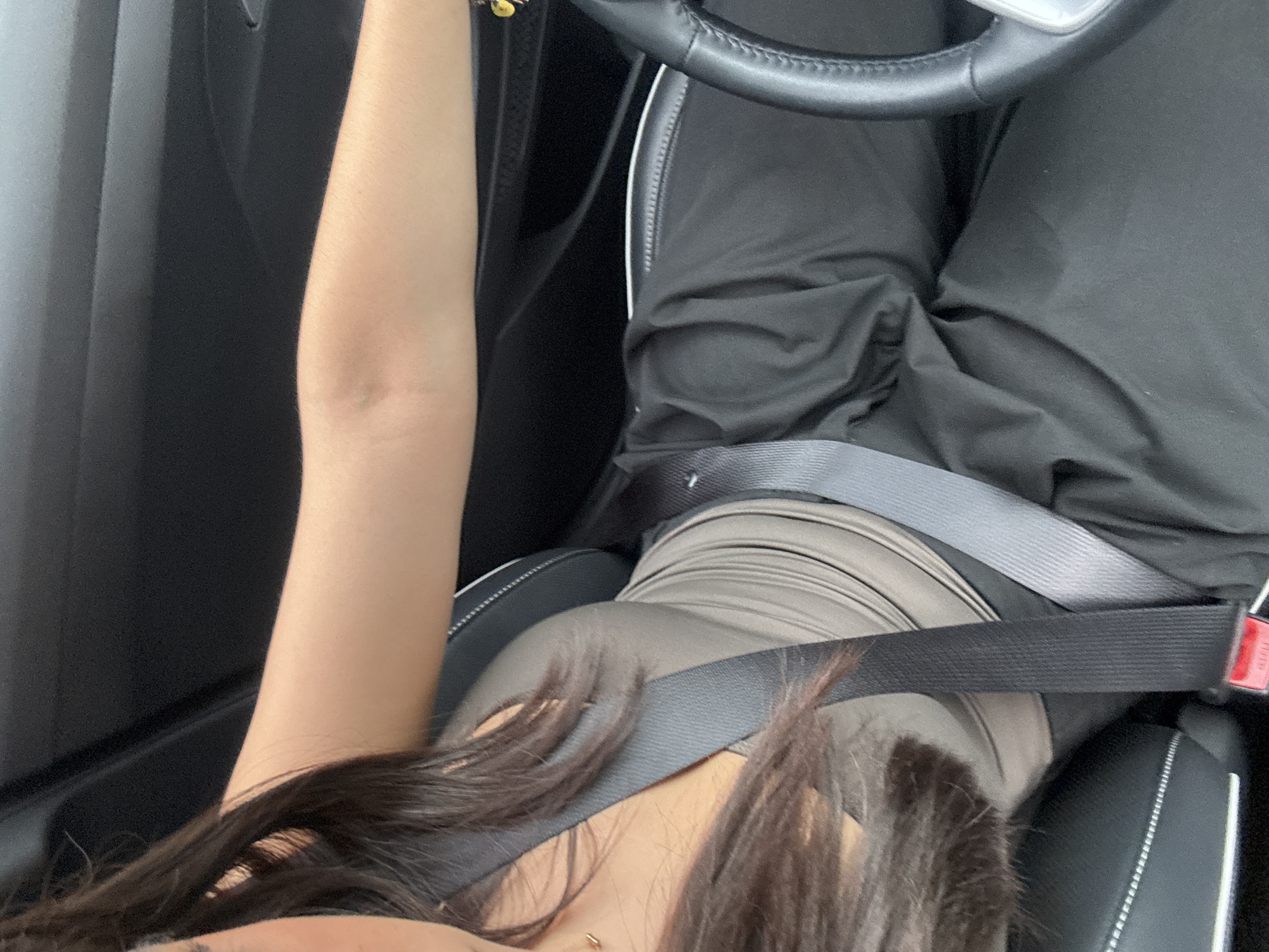 A seated person is wearing black pants and a tan top inside of a car. A seat belt is worn. The interior of the car is black. The person is holding the steering wheel.
The location is not identifiable as being in the city of Puebla, Mexico.