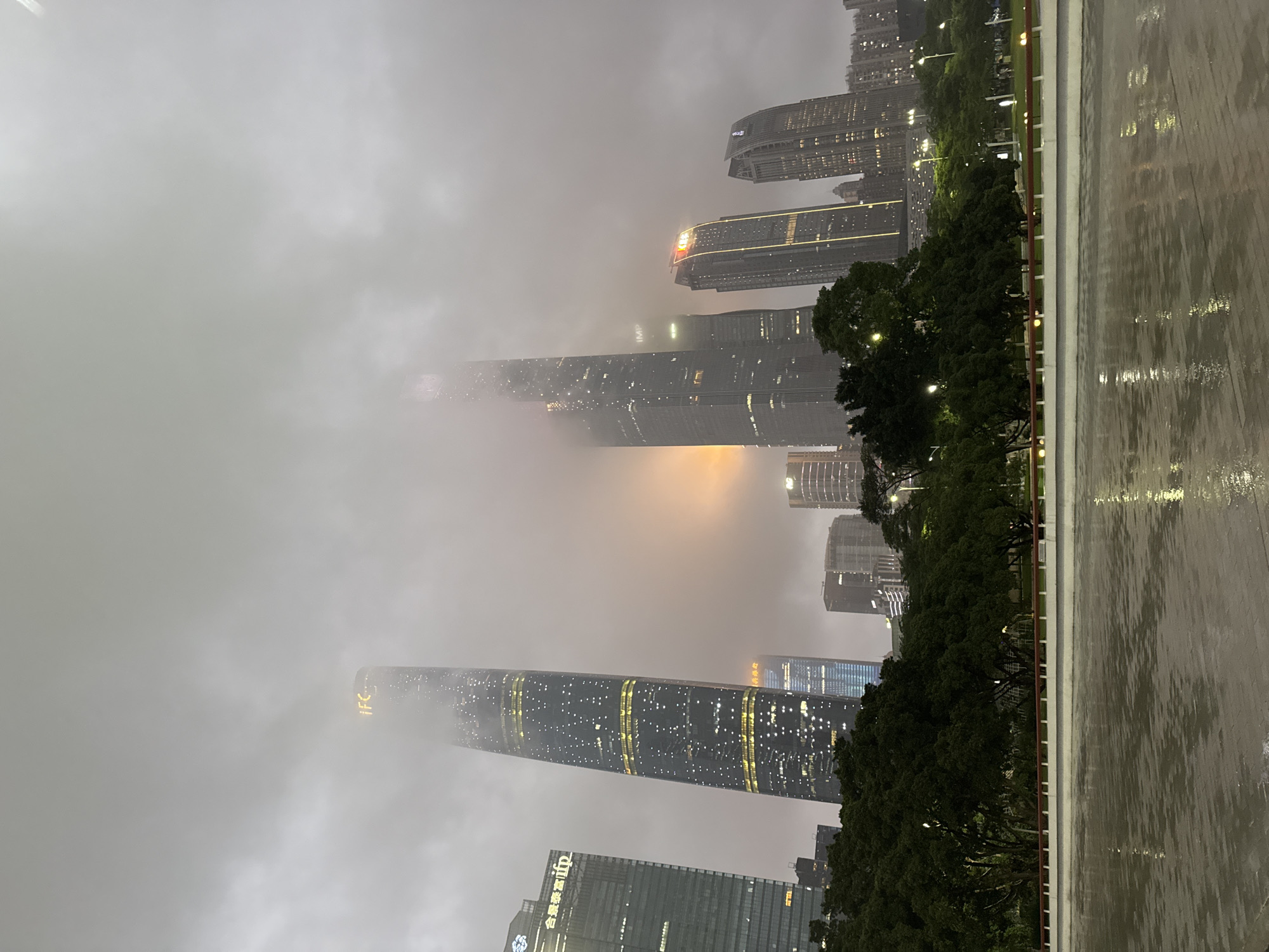 The image depicts a skyline of high-rise buildings obscured by heavy fog in what appears to be a wet environment. The location is identified as the Tianhe District in China, recognized by the presence of notable skyscrapers like the IFC tower. These buildings are partially illuminated with lights visible through the fog. In the foreground, a wet, reflective surface suggests recent or ongoing rain. Trees and some streetlights are also visible along the edge of the image. The sky is overcast, and the general atmosphere is muted due to the thick fog.