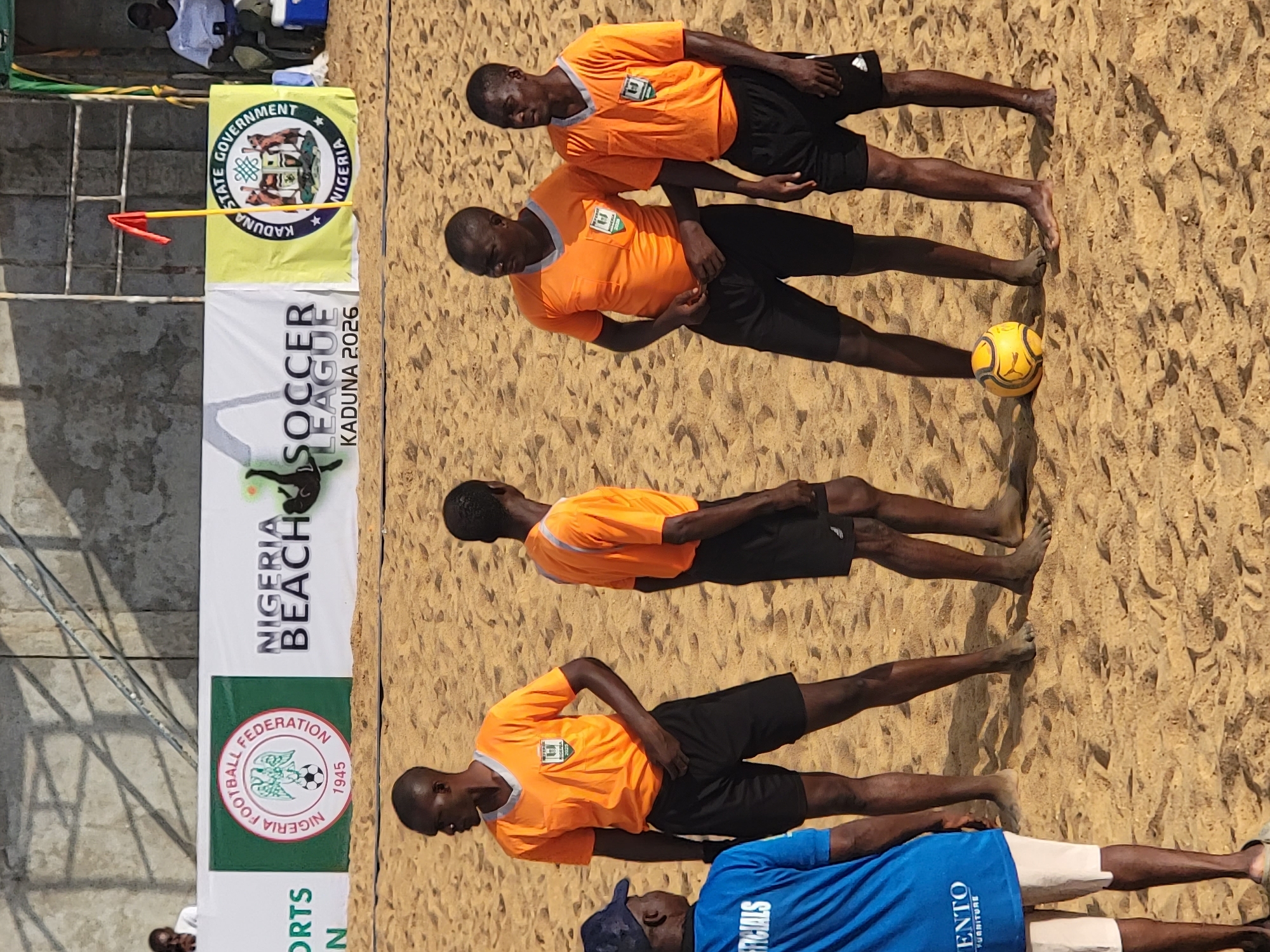 The image depicts a beach soccer scene, likely in Unguwar Sarki, Nigeria, as suggested by the "Kaduna 2026" banner, referencing a soccer league event. Four men in orange jerseys and black shorts, presumably beach soccer players or officials, are standing barefoot on the sand. One man is wearing a blue shirt, with the partial word "OFFICIALS" on it. A yellow soccer ball is on the sand. A banner with "Nigeria Beach Soccer League Kaduna 2026" is visible. The Kaduna State Government flag is also present. The surface is sand, typical of a beach soccer field.