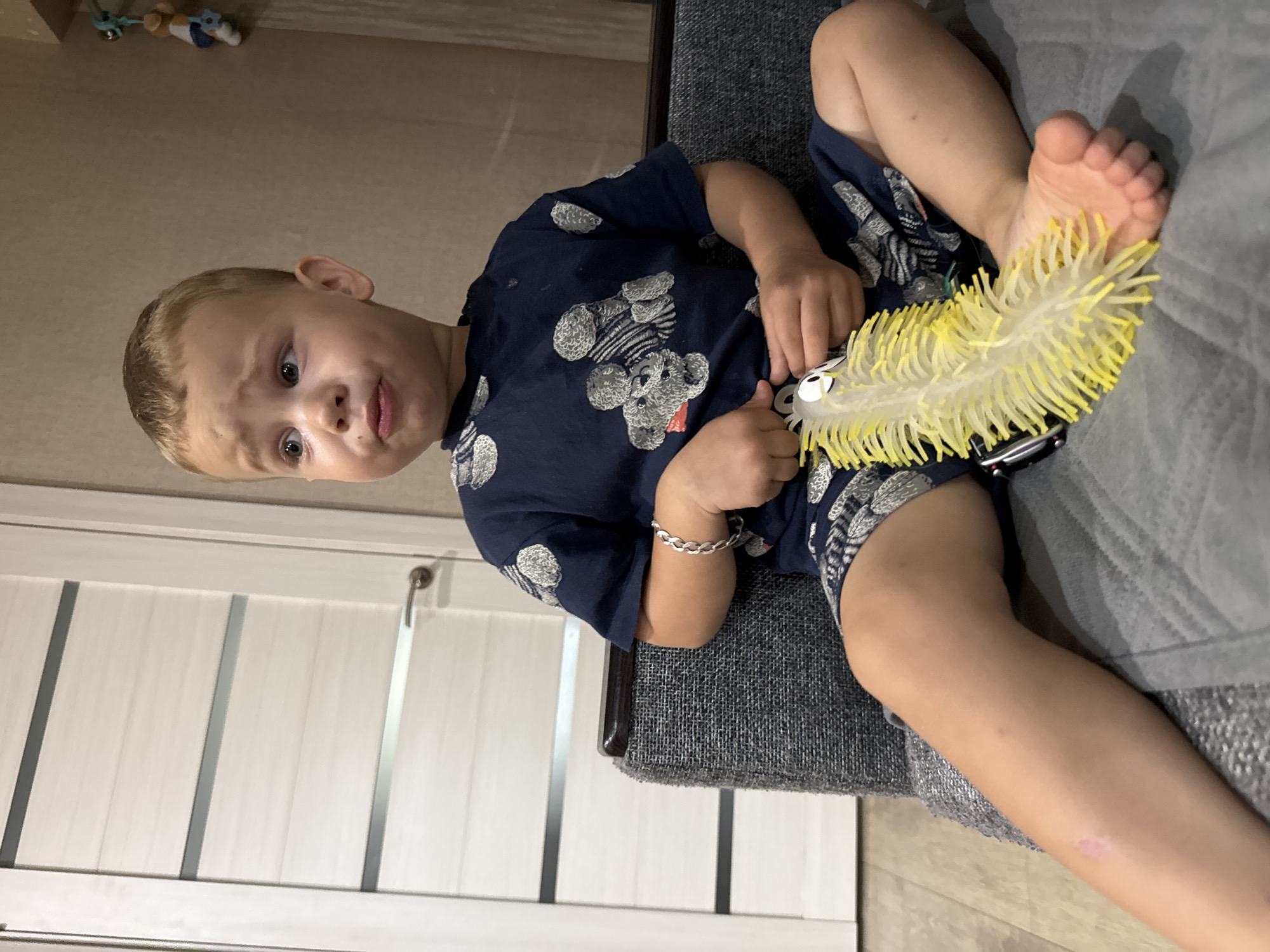 A young Caucasian boy with blond hair sits on a grey sofa, holding a yellow, spiky toy worm. He wears a dark blue t-shirt with a grey teddy bear pattern and matching shorts. The boy is looking to his right with a slightly pouted expression. To his left is a white door with horizontal grey stripes and a silver handle. The background consists of a plain beige wall and a part of a hanging mobile. The scene appears to be indoors, likely in a residential setting in Rusanovo, Russia.