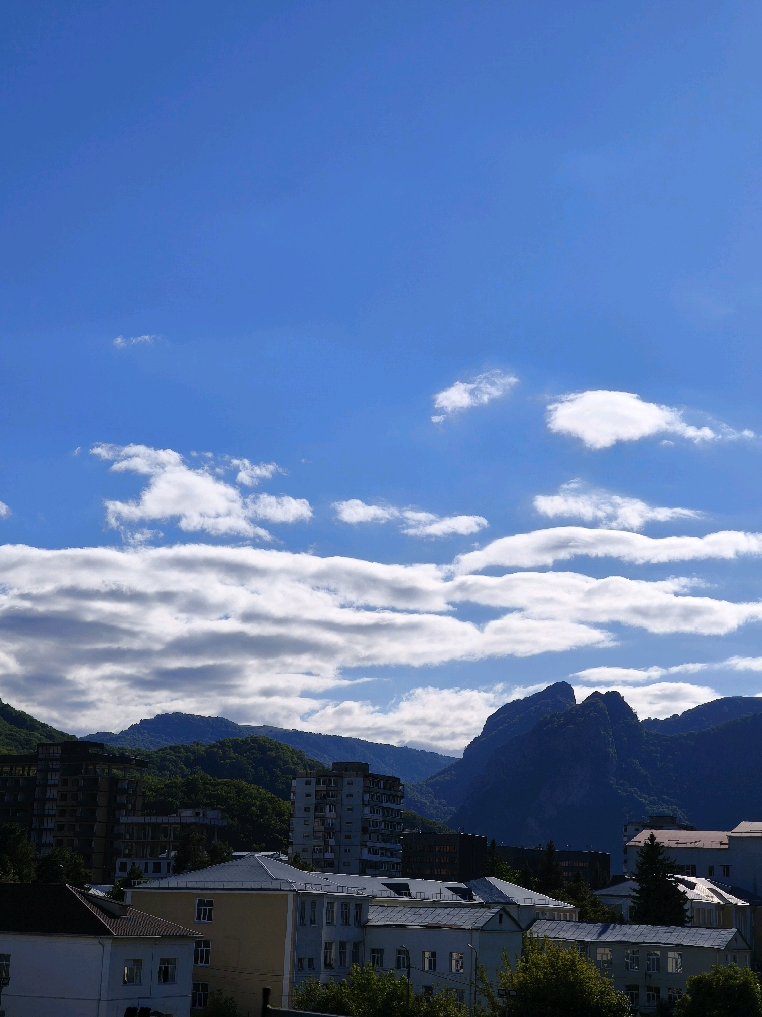 The image captures a view of Karachayevsk, Russia, featuring a residential area in the foreground and a mountain range in the background. The sky is a clear blue with scattered white clouds. Several multi-story buildings, typical of urban residential areas, are visible. These buildings vary in architectural style and color, with some appearing older and others more modern. Trees and greenery are interspersed between the buildings, suggesting a moderate level of vegetation within the city. The mountain range behind the city is covered in dense green foliage, indicating a forested or wooded landscape. The peaks of the mountains are rugged and appear to be a significant geological feature of the region. The overall scene depicts a blend of urban development and natural landscape.