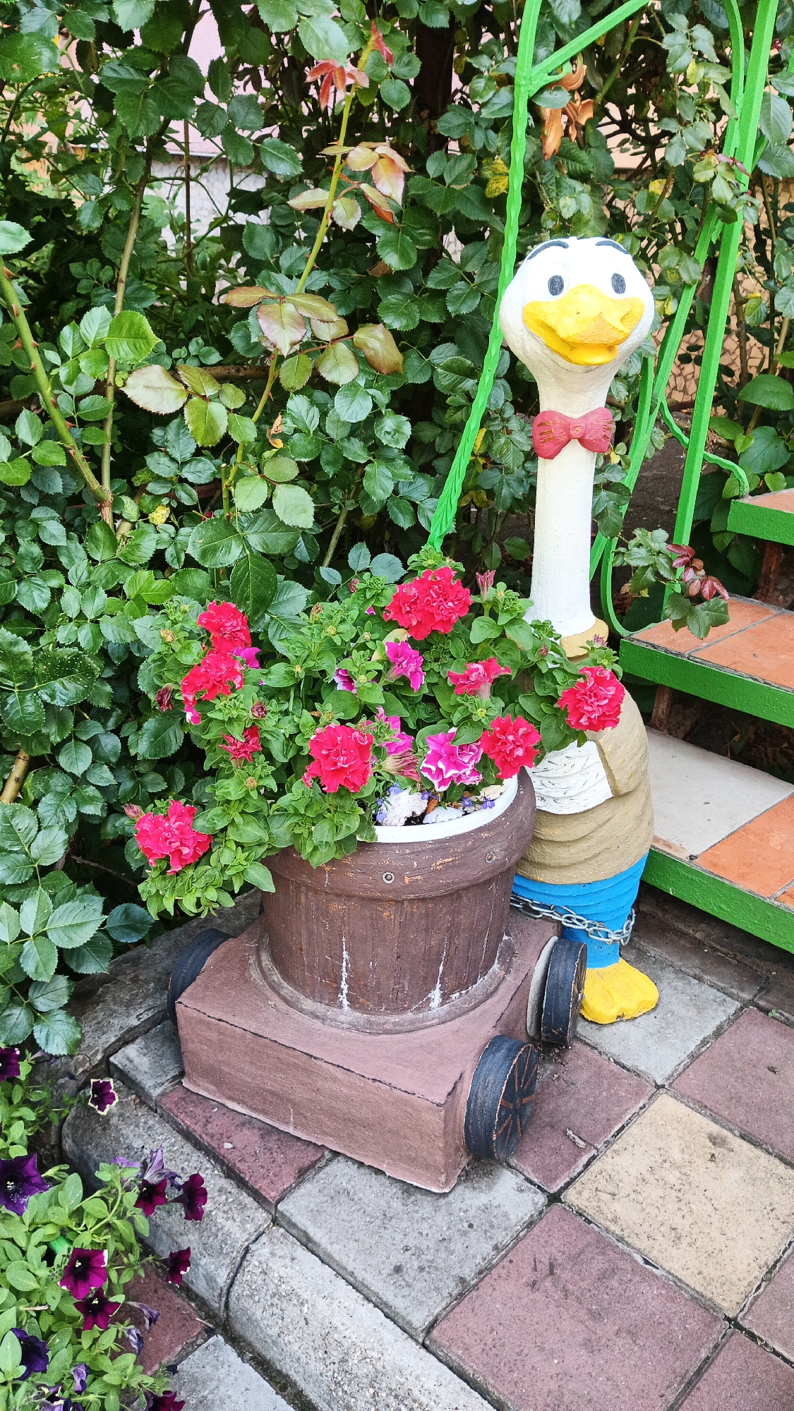 In Kryvyi Rih, Ukraine, a garden scene features a decorative ceramic goose figure with a red bow tie, standing next to a brown pot overflowing with vibrant pink and red petunias. The goose figure is painted with a white neck and head, a yellow beak, and black eyes. It wears a blue lower body and yellow shoes. The pot is on a wheeled base, also decorated in brown. Behind the flower pot and goose, a lush green rose bush with some new growth is visible. To the right, a green metal railing leads up a set of orange and brown tiled stairs. The ground is paved with interlocking bricks in shades of grey, red, and beige. In the lower left corner, a cluster of deep purple petunias adds another splash of color. The scene appears to be an outdoor decoration in a garden or yard.