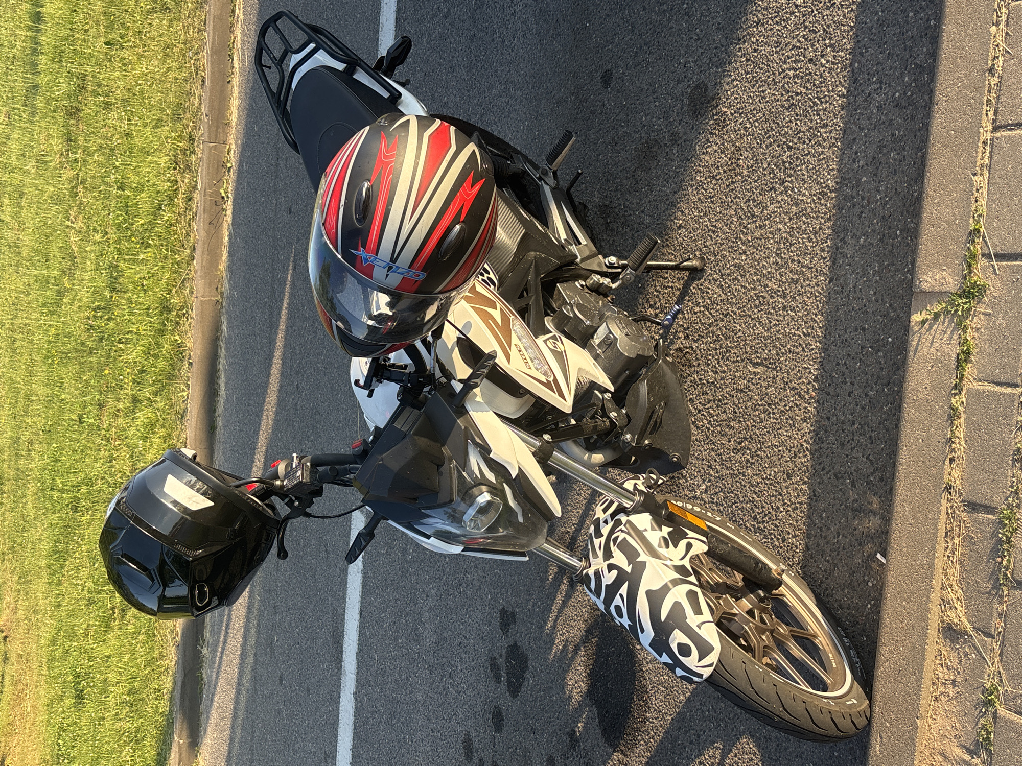 The image shows a motorcycle parked on an asphalt surface, possibly a road or parking lot, next to a grassy area. Two motorcycle helmets are visible; one is placed on the seat of the motorcycle, and another is resting on the handlebars. The motorcycle is predominantly white and black with some red accents on the red and white helmet. The front fender of the motorcycle features a distinctive black and white graphic pattern. The scene is illuminated by sunlight, suggesting it is daytime, likely late afternoon or early evening due to the long shadows cast on the asphalt. The grass appears dry, and there is a concrete curb separating the asphalt from the grass. No people are present in the image. There is some text visible on the red and white helmet, reading "KENZO," and on the side of the motorcycle, but it is mostly illegible.