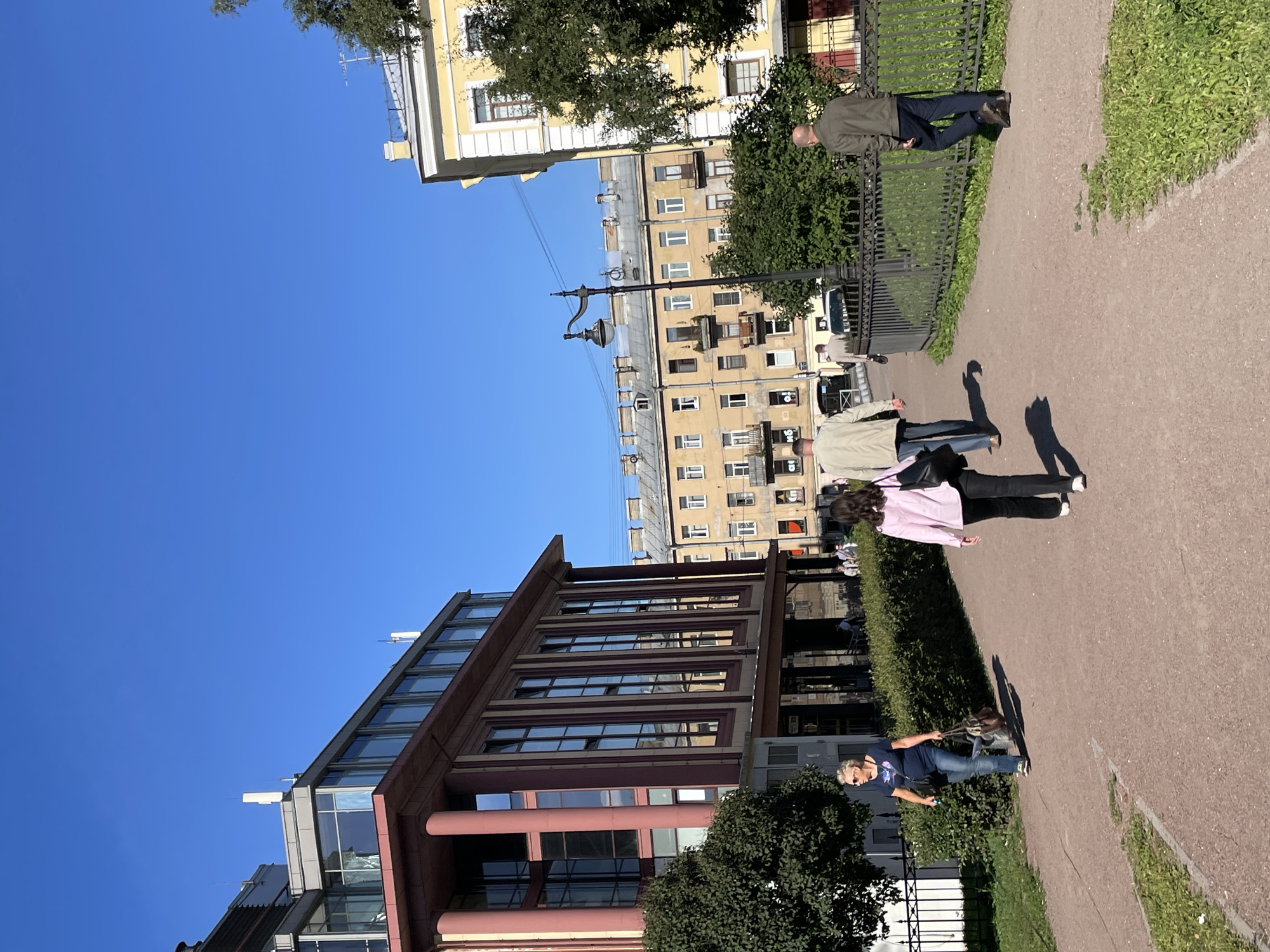 In Saint Petersburg, Russia, on a bright, clear day, a paved pathway curves through a park area bordered by grass and manicured hedges. On the left, a contemporary building with a reddish-brown facade and large glass windows stands prominently. To the right, a yellow, multi-story building with a traditional architectural style is visible. Several individuals are walking along the pathway. In the foreground, a woman with blonde hair, wearing sunglasses, a blue t-shirt, and jeans, walks to the left carrying a brown bag. Further down the path, a woman in a pink jacket and black pants walks away from the camera, followed by two men in casual attire, also walking away. In the middle ground, a streetlamp with a decorative design stands near the pathway. The background reveals more residential buildings with visible windows and balconies, indicative of a city environment. The overall scene depicts a pleasant urban park setting on a sunny afternoon.