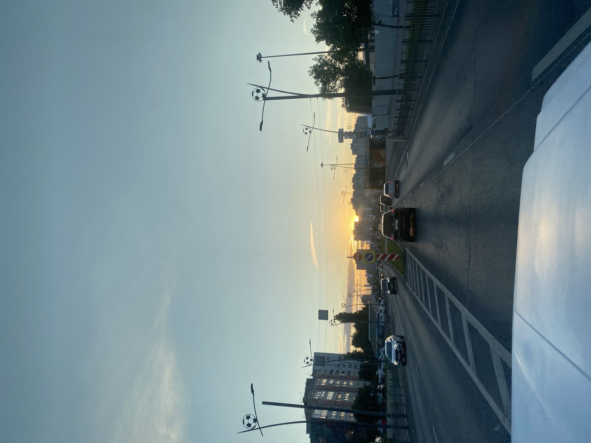 The image captures a view from a vehicle looking forward on a multi-lane road in Nizhny Novgorod, Russia, during sunset. The sky is a gradient of light blue and pale orange, with scattered clouds. The sun is visible on the horizon, casting a warm glow. Along the side of the road, there are lampposts topped with soccer balls, suggesting a thematic design possibly related to a sporting event or venue. Residential buildings, trees, and other streetlights are visible on the left and right sides of the road. Several cars are driving in the same direction as the viewer, and there are traffic signs and road markings. The foreground shows the hood of the vehicle from which the image is taken.