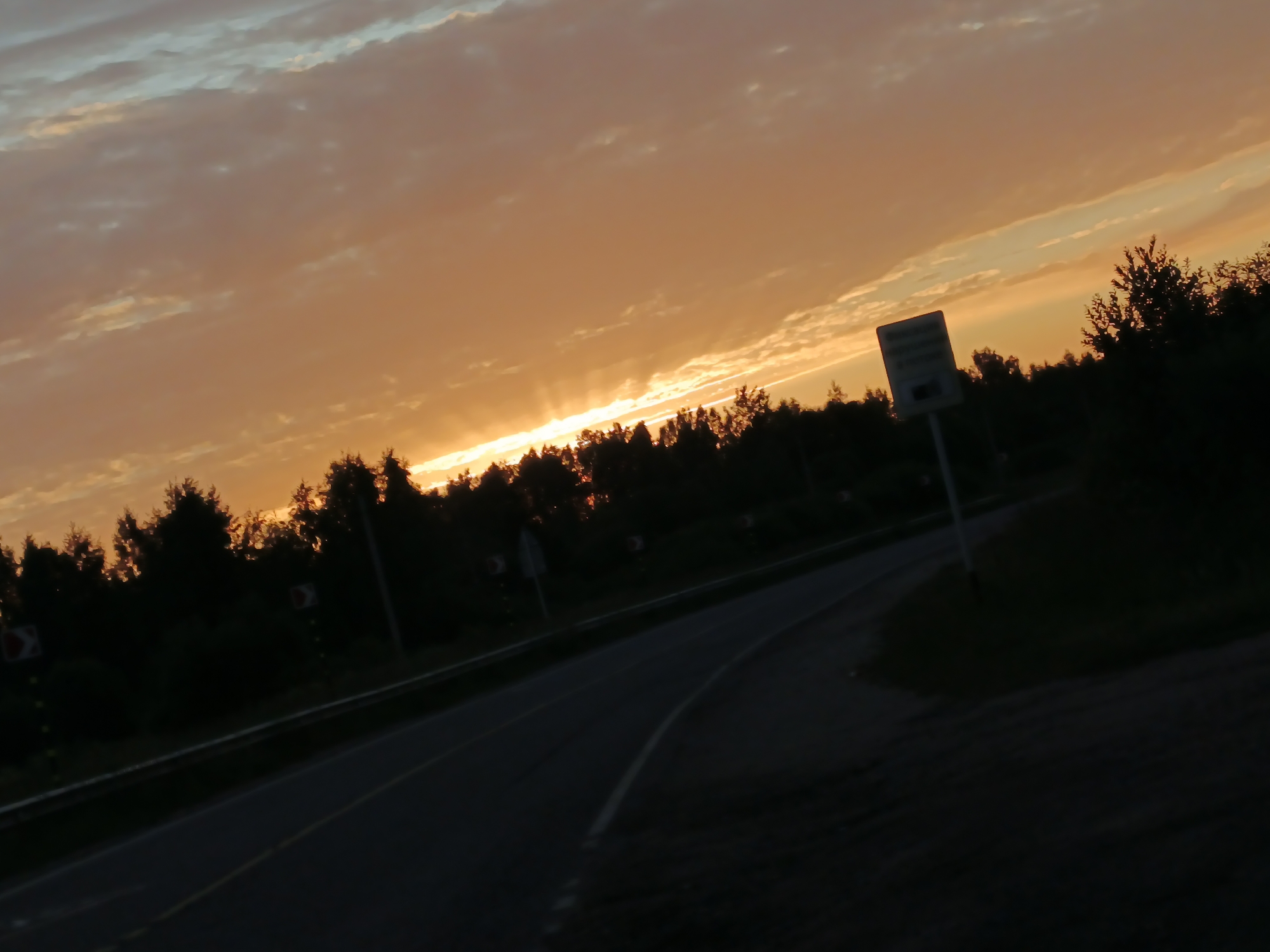The image depicts a road at dusk, with the sky painted in hues of orange and yellow from the setting sun. Silhouetted trees line the right side of the road, and there are traffic signs visible. The road itself is paved, with a white line marking its edge. The scene is set in Воскресенское, Russia.