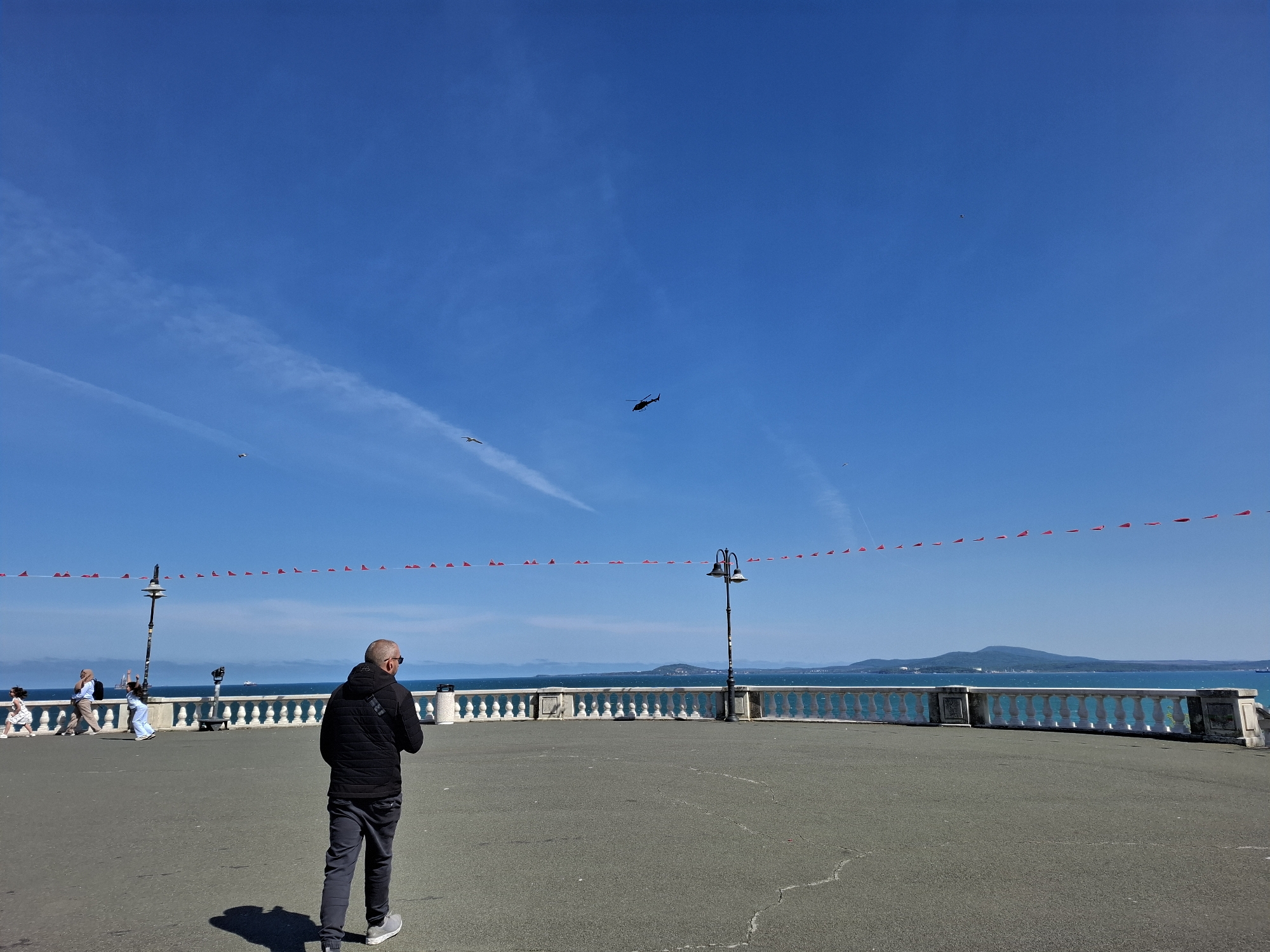 The image shows a partially-filled seaside promenade in Burgas, Bulgaria, on a sunny day. A man in a black jacket and dark pants walks away from the viewer. Other people are walking and playing to the left. A white stone balustrade runs along the sea. A row of red pennants hangs above. In the clear blue sky, a helicopter and several birds are visible. Two streetlights stand on the promenade. In the background, the sea meets a distant mountainous landmass.