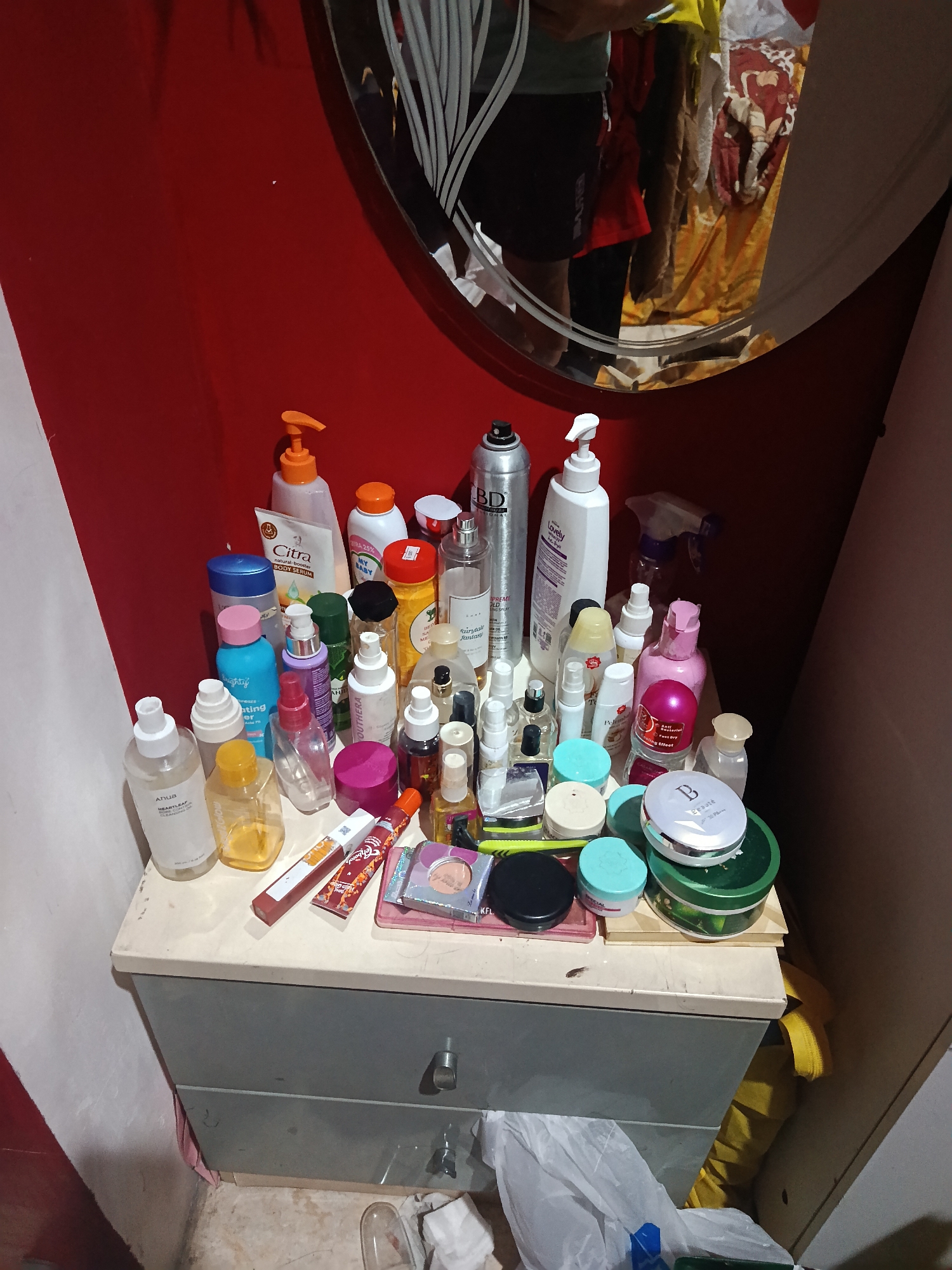 The image shows a dresser with various cosmetic products on top. The dresser is dark gray with two drawers. The top of the dresser is a light gray surface where the products are arranged. The products include bottles of lotion, sprays, powders, and other beauty items. Among the visible items are a "Citra natural white body serum" and a "BD PROFESSIONAL" hair spray. A mirror is visible in the background, reflecting a person wearing dark shorts and a yellow shirt, as well as some clothing items. The wall behind the dresser is painted red. The floor around the dresser is tiled. A white plastic bag and a blue bottle are visible on the floor in the lower right portion of the image. The scene appears to be an indoor setting, possibly a bedroom or dressing area.