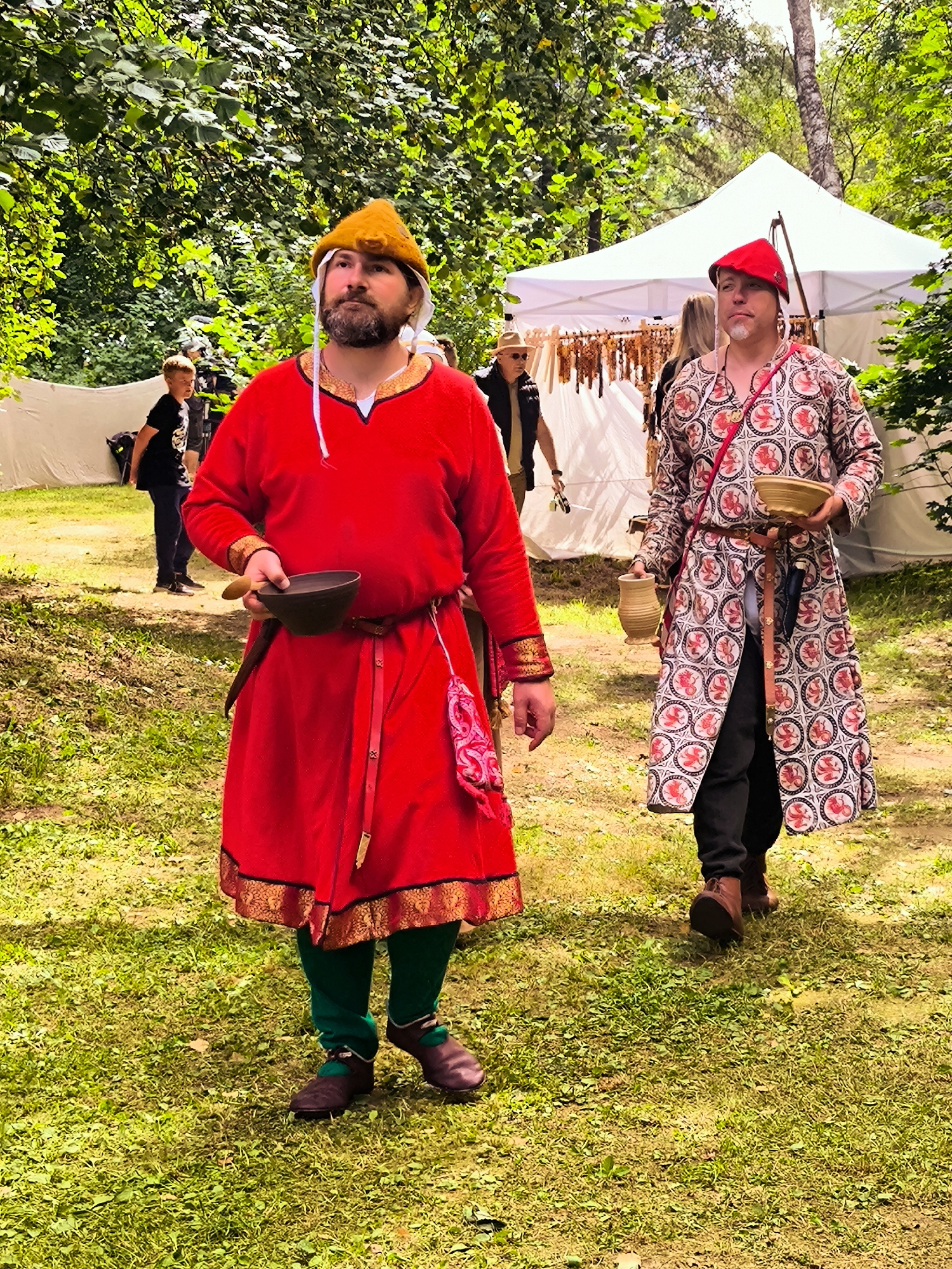 Two men in historical reenactment costumes are walking on a grassy area with trees in the background. The man in the foreground, on the left, wears a bright red tunic with gold trim and a yellow conical hat. He has a beard and is holding a dark bowl with a wooden spoon. He is also wearing green leggings and brown shoes. The man in the background, on the right, wears a patterned long coat with red and white circular designs, and a red cap. He is holding a small clay jug and a shallow wooden bowl. Behind them, a white tent is visible, and a young boy in a black t-shirt stands in the distance. This scene appears to be from an outdoor event or festival, possibly in Tērvete, Latvia, given the natural, wooded environment.