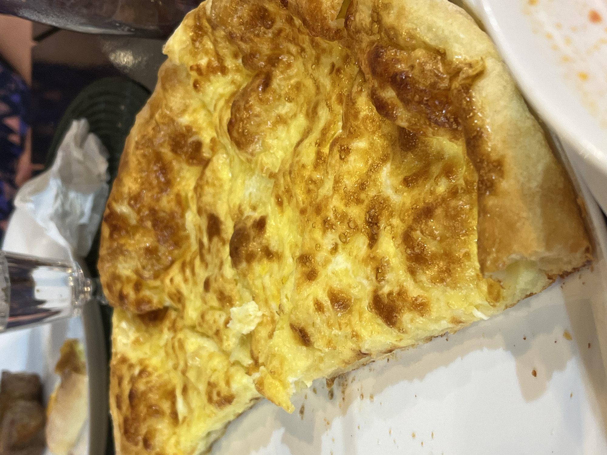 A close-up shot of a khachapuri, a Georgian cheese-filled bread, rests on a white plate. The pastry is golden brown and appears to be filled with melted cheese. In the background, out of focus, a glass of water, a napkin, and a dark bottle are visible, suggesting a dining setting. The image is taken from a high angle, with the khachapuri filling most of the frame. The location is identified as Kobuleti, Georgia.