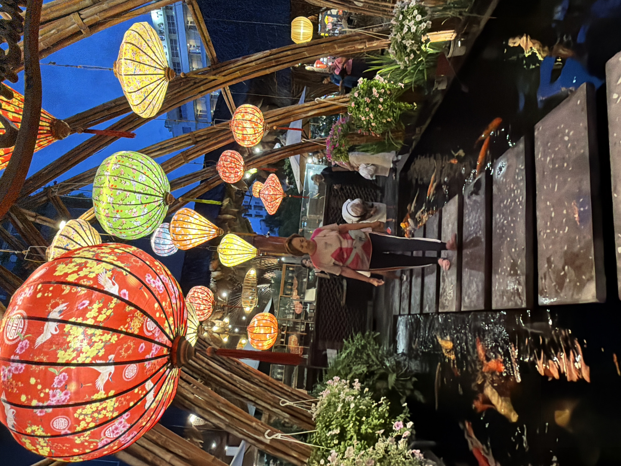This is a photograph of an outdoor dining area at night in Nha Trang, Vietnam, decorated with numerous traditional Vietnamese lanterns of various colors, shapes, and sizes, suspended from a bamboo structure. The lanterns are illuminated, casting a warm glow. In the foreground, a woman with short brown hair, wearing a pink t-shirt and dark pants, walks across a stone path that bridges a koi pond. The pond contains several koi fish swimming. Lush green plants with small pink and white flowers flank the path and pond. In the background, other diners and staff can be seen, and a building with illuminated windows is visible. The sky is dark blue, indicating it is evening.