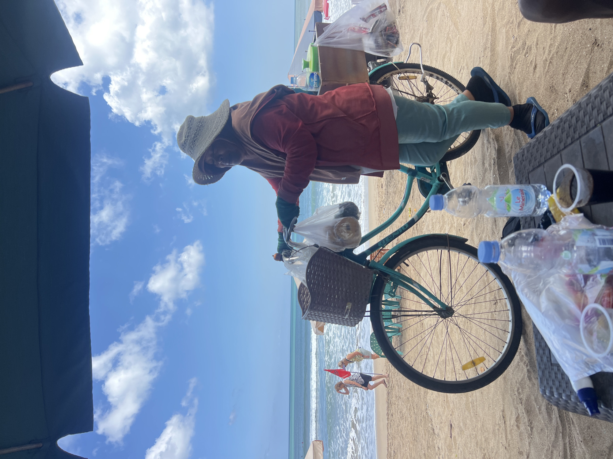 A person wearing a large straw hat, a brown hijab, a red jacket, and light green pants stands on a sandy beach next to a teal bicycle. The bicycle has a brown basket on the front with plastic bags of goods, and a wooden box behind the seat with more items and a water bottle. The person is holding a plastic bag in their left hand. In the background, the ocean is visible with waves, and two women are walking on the beach. Beach umbrellas and chairs are also present. A table in the foreground holds a plastic bag, a cup of dark liquid, and two water bottles. The sky is blue with scattered clouds. The scene is set on Kuta Beach in Bali, Indonesia.