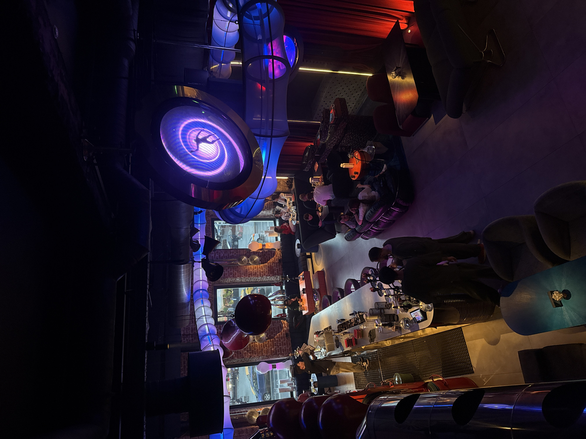 The image shows the inside of the chain restaurant, "Didu," located in Moscow, Russia. The restaurant has an industrial-chic decor. Round tables with black and orange tops are surrounded by customers sitting on chairs. Above the tables, there is a lit, round decorative light fixture displaying a person in a twisting motion. In the background, a bar is visible, stocked with glasses, alcohol, and a bartender. The location appears to be on a busy street with car traffic.