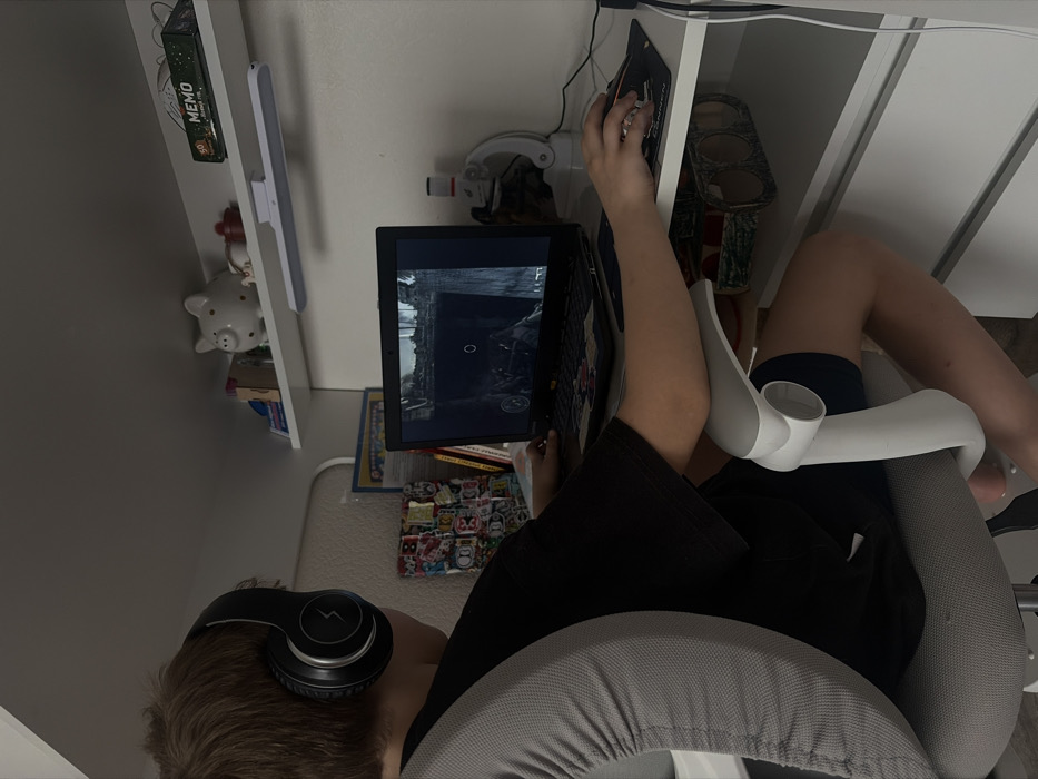 An individual with light brown hair, wearing black over-ear headphones with a lightning bolt logo, a black and grey short-sleeved shirt, and black shorts, is seated at a white desk. The person is positioned with their back to the camera, engaged in playing a video game on an open black laptop. The laptop screen displays a dark, possibly industrial or ruined environment from a first-person perspective, featuring a circular reticle. The individual's left hand rests on the laptop keyboard, while the right hand operates a black computer mouse on a dark mousepad. To the right of the laptop, a white and red instrument resembling a small microscope is visible. Above the desk, a white floating shelf holds a white ceramic piggy bank with black spots, a small red lidded container, a green box labeled "MEMO," and other small items. A white LED light bar is mounted beneath this shelf. The person is seated on a grey ergonomic office chair with light grey armrests. The walls are white. This scene is located in Krasnaya Zarya, Russia.
