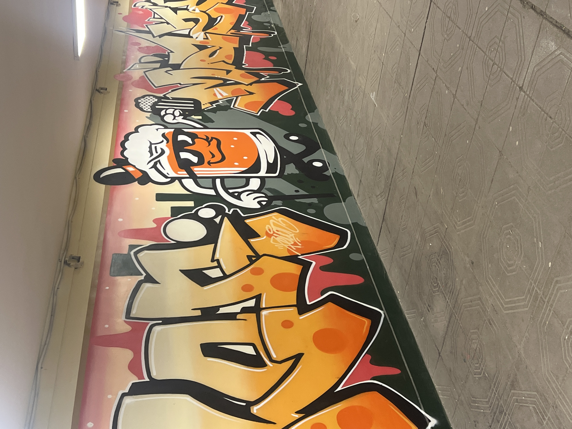 The image shows graffiti art on a wall next to a sidewalk. The graffiti is primarily orange and white with black outlines. A cartoon drawing of a beer mug with a face and arms is featured prominently. The sidewalk is made of gray tiles with a geometric pattern. The graffiti and sidewalk are likely located in an underpass. This is located in Sofia, Bulgaria.