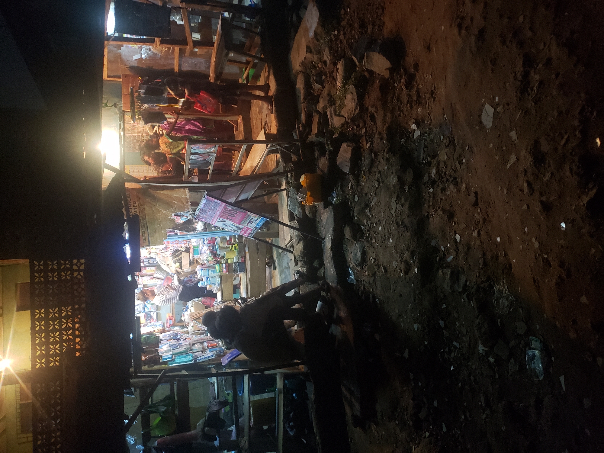 The image depicts a night scene in Alalubosa, Nigeria. A small retail shop is illuminated by bright overhead lights, revealing a variety of merchandise on shelves. Several people are inside the shop, with one person seemingly engaged in a transaction. Outside the shop, the ground is uneven with dirt and rocks. Additional objects are present in the vicinity of the shop, including what appear to be containers or packages. The foreground is dark, suggesting limited ambient lighting outside the immediate vicinity of the shop.