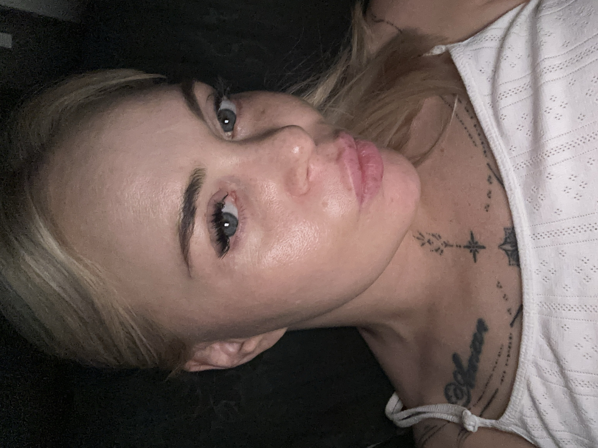 A close-up, selfie-style portrait depicts a woman with blonde hair and blue-grey eyes, positioned in a dark, indistinct environment. Her face is directed towards the camera, maintaining a direct gaze, with her lips pursed in a "kissing" or "pout" expression. Visible eyelashes appear long and full. Her skin exhibits a slight sheen, particularly on the forehead and cheeks, with visible texture. Faint lines are present on her forehead. She wears a white tank top featuring thin spaghetti straps and a patterned fabric with small, repeating diamond-shaped perforations. Several tattoos are visible: on the left side of her neck, a vertical design composed of leaf-like elements culminating in a star symbol, with a partial geometric pattern below it; on her upper chest, above the left breast, the word "Amor" in script lettering, accompanied by a line of small dots/dashes; and partially visible on the right side of her chest, a star or compass rose design. The background is uniformly dark and lacks any identifiable features. The location is Unknown, Unknown.