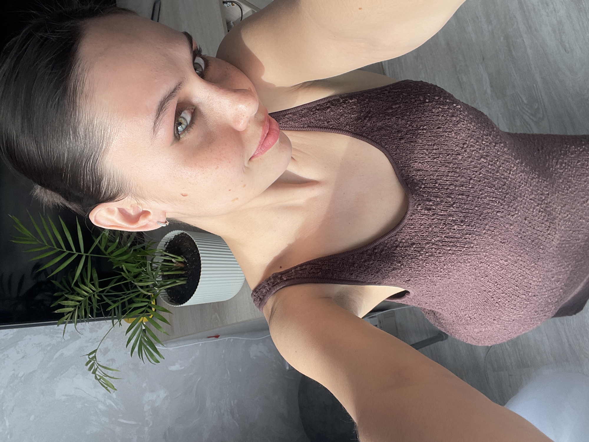 A selfie shot features a woman with dark hair pulled back, fair skin with freckles, and green eyes, wearing a brown tank top. She is positioned in the foreground, looking directly at the camera with a slight upward tilt. To her left, a potted plant with green leaves stands next to a television. The background consists of a light gray surface, possibly a table or floor. The location in the city of Izhevsk, Russia is not discernible from the image.