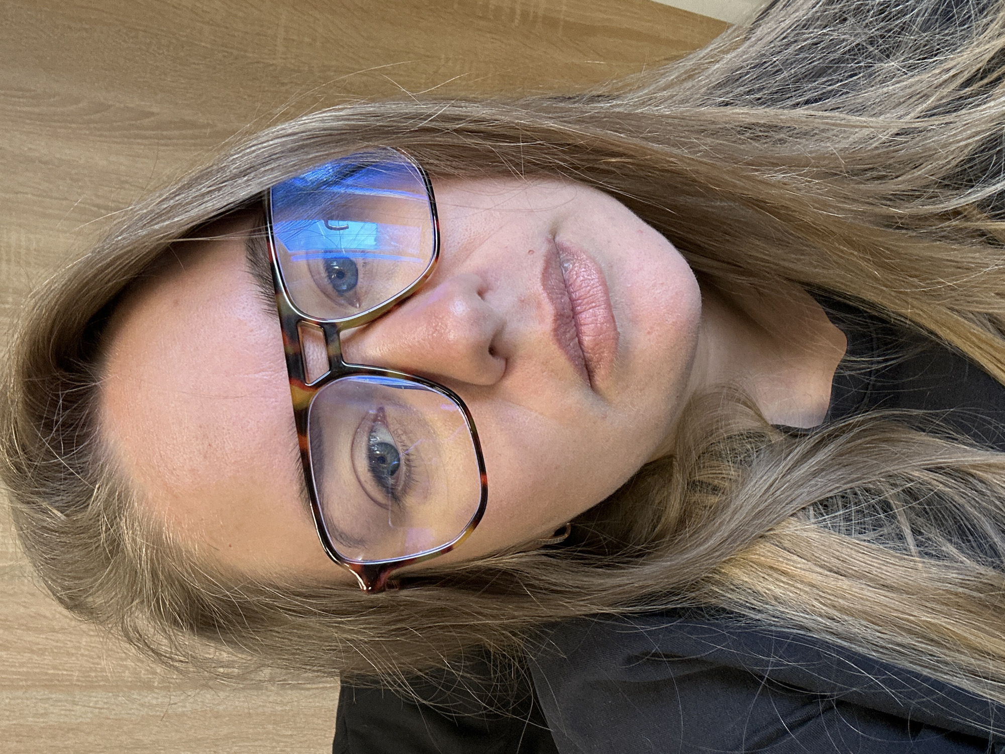 A woman with blonde hair and blue eyes wearing tortoiseshell glasses is photographed in a close-up, angled view. The background consists of a wooden surface and a black fabric garment. No specific details suggest the location is Ural'skoe, Russia; this cannot be confirmed visually.