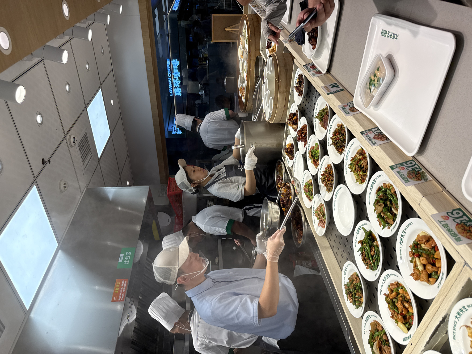 The image depicts a busy food service area, likely a cafeteria or restaurant, where customers select pre-prepared dishes from a counter. Several food service workers, wearing hairnets and some with face shields, are actively engaged in serving and preparing food. One man in the foreground, wearing a light blue shirt and a white chef's hat, is holding a ladle over a metal pot, seemingly serving a dish. Other workers are visible in the background, some tending to cooking stations.
The counter displays a variety of dishes served in white plates, arranged in rows. These dishes appear to be Asian cuisine, with visible ingredients like vegetables, meats, and possibly rice. Some plates have small labels with Chinese text. There are also stacked bowls and other serving utensils on the counter.
In the background, there's a sign that reads "LOWAN COFFEE" in English and Chinese characters, suggesting this area might be within or adjacent to a coffee shop. The overall environment is well-lit, with overhead fluorescent lights and spotlights. The scene captures a typical bustling food service operation with customers being served. The presence of Chinese text on signs and food labels indicates the location is in China. The hint image showing "天河区" (Tianhe District) suggests the specific city area.