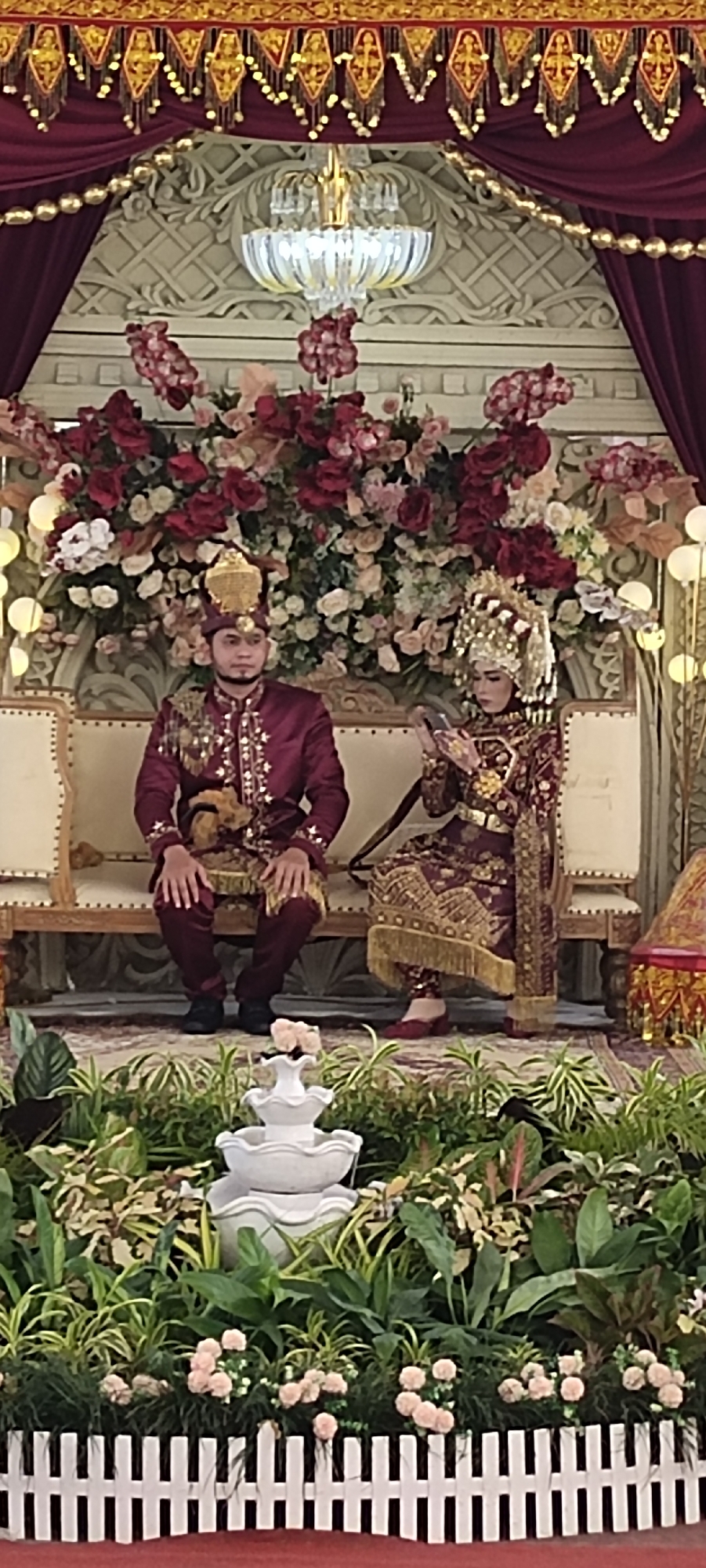A photograph depicts a wedding scene. A man and a woman, presumed to be the bride and groom, are seated on a decorative bench. They are adorned in traditional formal attire, colored in shades of burgundy and gold. The man is seated to the left, and the woman to the right.
Behind the couple is a backdrop of red and white flowers and greenery, with a chandelier visible above. Above that, there is elaborate golden decoration and red curtains.
In the foreground, a white tiered fountain is surrounded by various plants and flowers, all contained by a white picket fence.
The location is identified as Unknown, Unknown. The overall scene suggests a formal, traditional wedding ceremony.