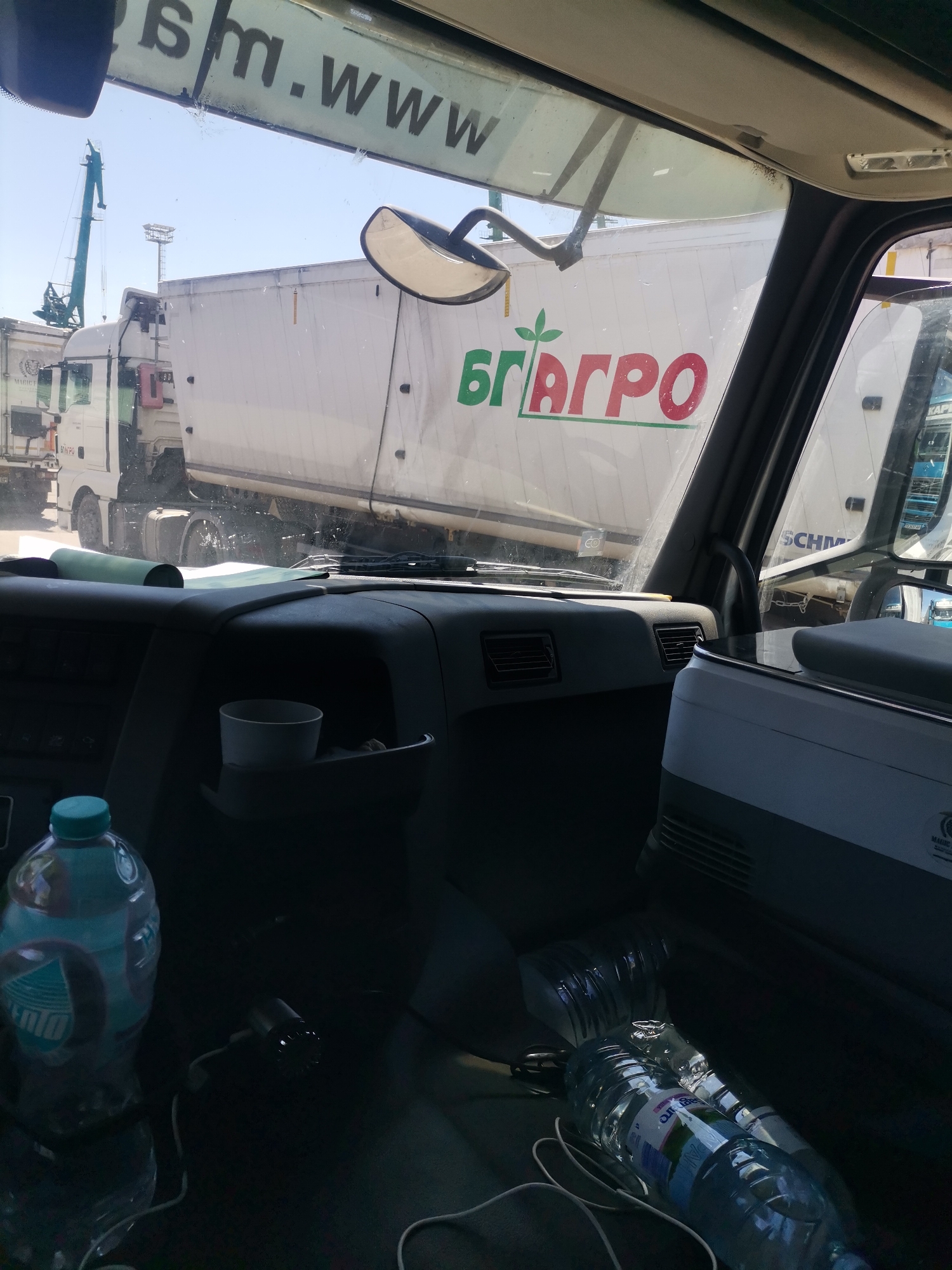 The image appears to be taken from inside the cab of a truck, possibly a semi-trailer truck. The view through the windshield shows other trucks, including one with the logo "БГ АГРО" in green and red lettering on a white trailer. There is a crane visible in the background, suggesting a port or industrial area. The interior of the truck cab contains water bottles, one with a blue label, and a cup in a holder. The "m.www" lettering is partially visible on the top of the windshield, obscuring other text. Based on the presence of the Cyrillic alphabet and the "БГ АГРО" logo, it is very likely that this image was taken in Bulgaria. The port or industrial area suggests that the location is in the city of Varna, Bulgaria, a port city on the Black Sea.