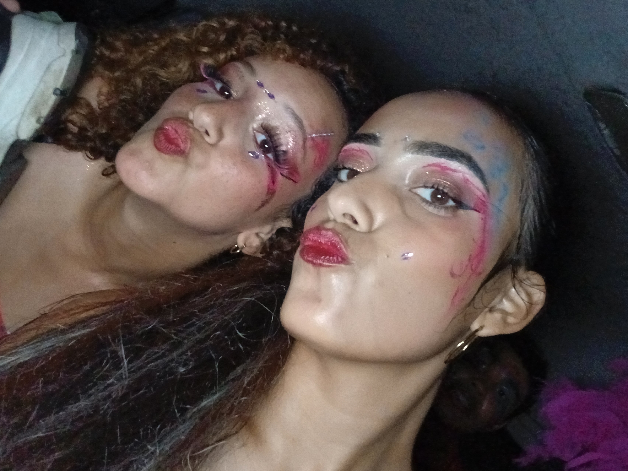 Two women with elaborate makeup, including heavy eye makeup, lip color, and decorative accents, are posing closely together. The woman on the left has curly hair, while the other has straight, dark hair. The scene has low lighting, and there is a dark figure in the background partially visible near the ear of the woman on the right. The specific location in Playa El Agua, Venezuela, cannot be determined from the visual information.
