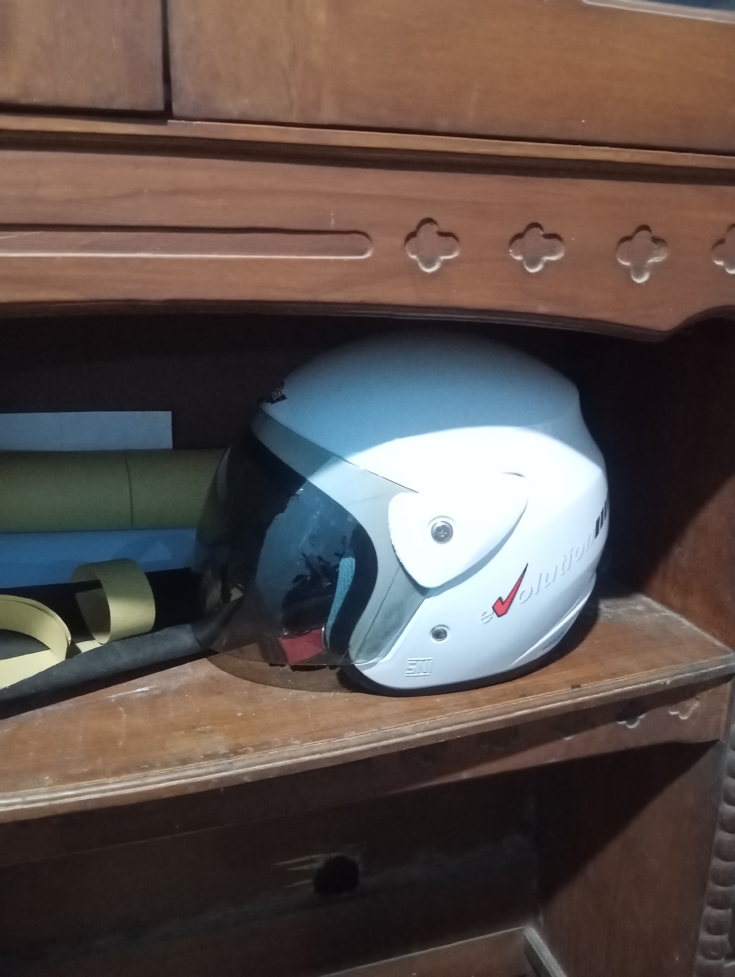 A white motorcycle helmet with a dark tinted visor is placed on a wooden shelf in what appears to be a piece of furniture with decorative carvings. The helmet has a red checkmark logo and text that reads "evolution" in black. To the left of the helmet are several rolls of paper or material, including one that is yellow and another blue. The shelf and furniture are made of dark wood. The image is likely taken indoors. Based on the context provided, the scene is located in Sudikampiran, Indonesia.