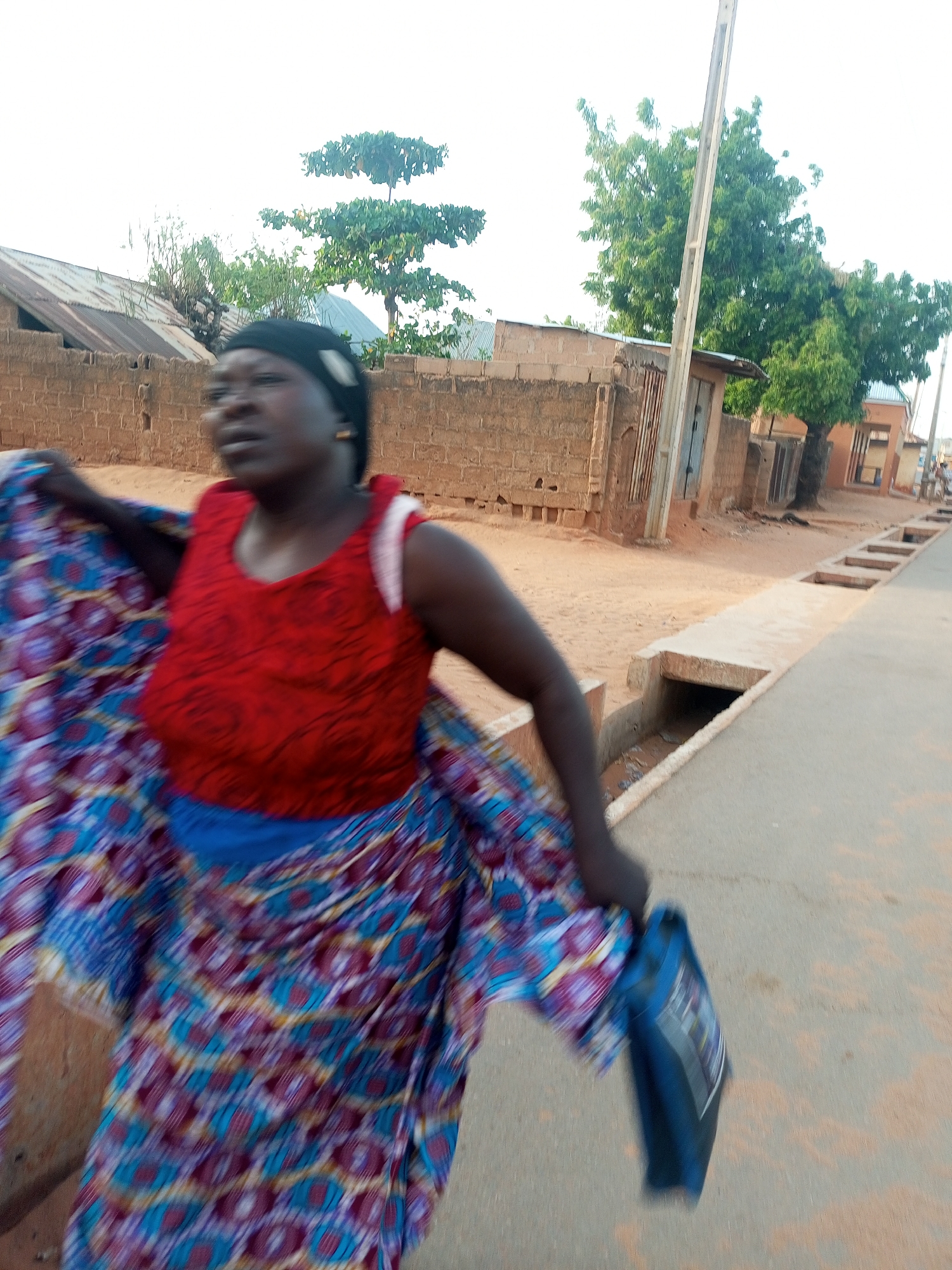A woman in a red sleeveless top and a patterned blue skirt is walking on a paved road. She is carrying a blue bag. The background includes brick buildings, trees, and a utility pole. The location is Tempure, Nigeria.