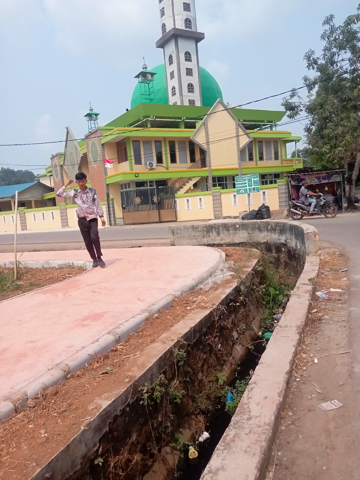 This is a medium shot, taken outdoors during daylight, featuring a young man standing on a newly constructed pink-colored walkway. The man, dressed in a patterned long-sleeved shirt and black pants, is looking towards the camera and has his right hand raised to his forehead in a gesture. Behind him, to the left, is a mosque with a green dome and a tall, white minaret. The mosque's facade is painted in yellow and lime green. To the right of the mosque, a person is riding a motorcycle on a paved road. A drainage ditch filled with water and some debris runs along the edge of the walkway and the road. The area appears to be under development, with exposed soil and construction materials visible. The overall setting suggests an urban or suburban environment in Batam, Indonesia, likely near a public building or a residential area.