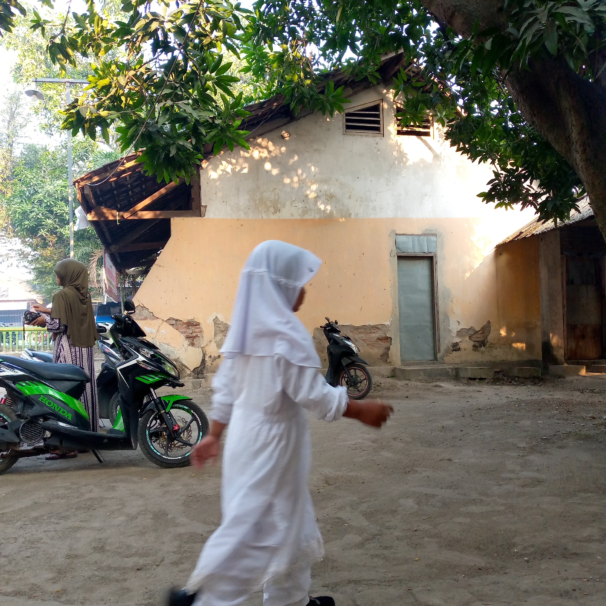 The image depicts an outdoor scene in Pacing, Indonesia, with a focus on a residential area featuring a partially dilapidated building. In the foreground, two individuals are present. One is a young person wearing a white hijab and a white garment, walking from left to right and out of frame. Their movement is blurred, indicating they are in motion. To the left, another person, likely an adult, is standing next to a green and black motorcycle. This individual wears a dark olive-green hijab and a patterned long-sleeved dress. They are holding a dark object in their hands. Two motorcycles are visible: a green and black one on the left, with the brand "HONDA" visible on its side, and a black motorcycle further to the right, partially obscured. The building in the background has an earthy-toned facade with visible signs of wear and tear, including exposed brickwork and peeling plaster. A tiled roof is partially visible, supported by wooden beams. Sunlight casts dappled shadows from the trees onto the building's wall. The ground is covered in dirt and gravel. Trees with lush green foliage overhang the scene, providing shade. The overall impression is that of a casual, everyday moment in a rural or semi-rural Indonesian setting.