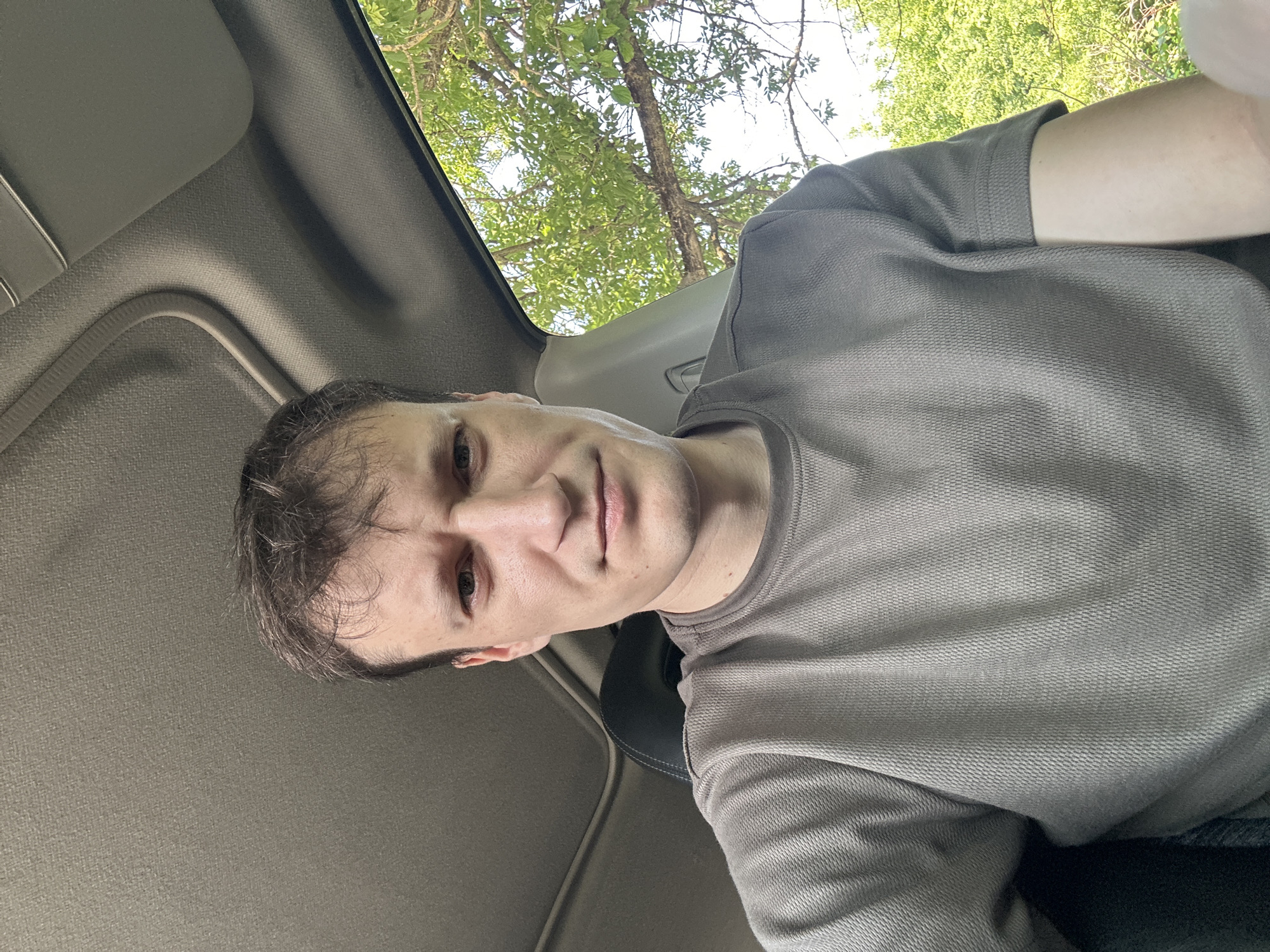 A man with short brown hair and fair skin is seated inside a vehicle. He is looking directly at the camera with a neutral expression. He is wearing a short-sleeved, textured grey t-shirt. The interior of the car, including the roof lining and a sun visor, is visible behind him. Outside the vehicle, through the window, lush green trees and foliage are visible under a bright sky, suggesting an outdoor, possibly rural or suburban, setting. The image appears to be a selfie taken from a slightly elevated angle. The location is not discernible from the image content.