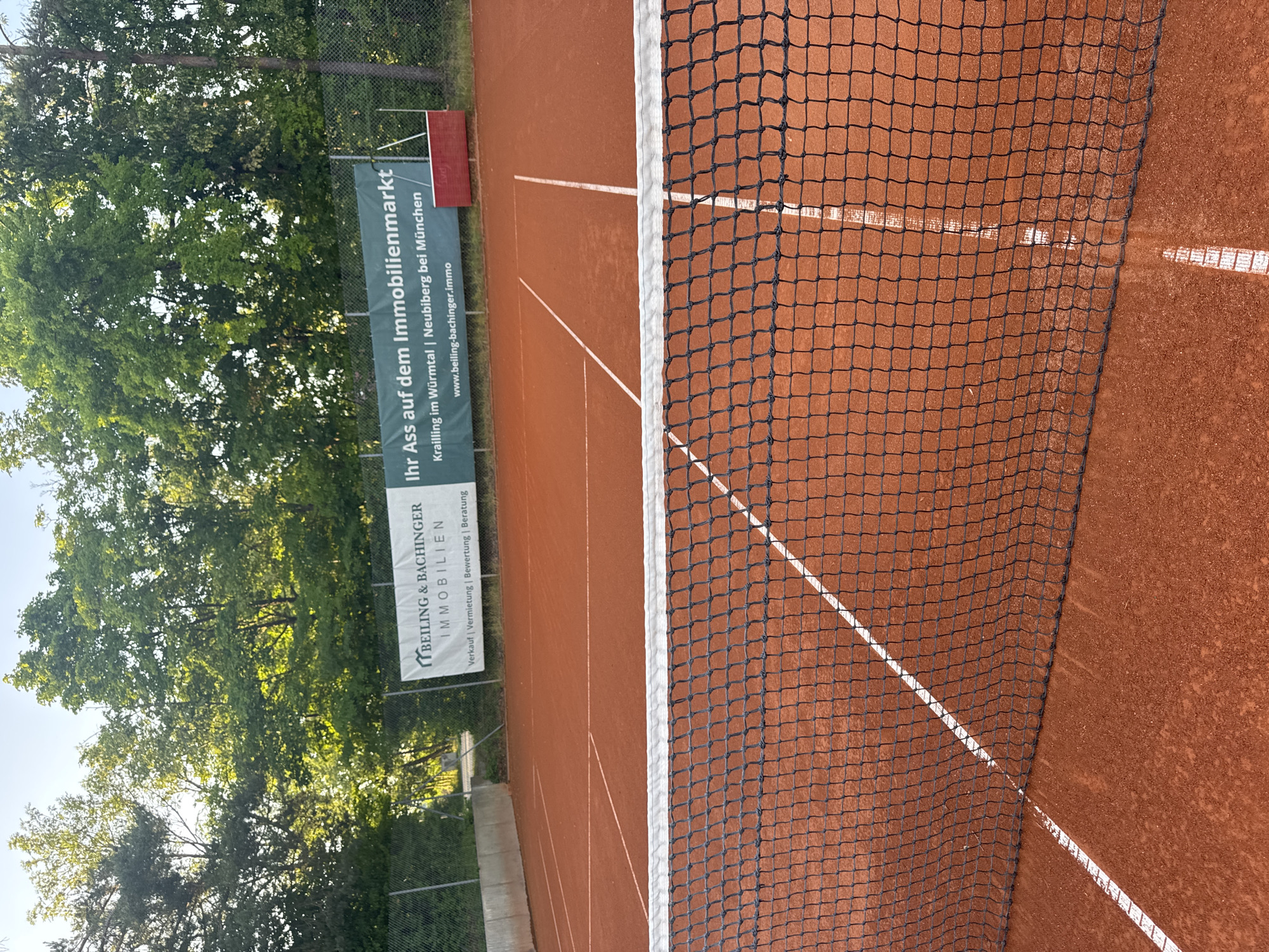 An outdoor clay tennis court is shown, with a net in the foreground. The court has white boundary lines. In the background, there is a fence and trees. A banner is attached to the fence, reading "Ihr Ass auf dem Immobilienmarkt" and "Krailling im Würmtal | Neubiberg bei München". The website "www.beiling-bachinger.immo" is also visible on the banner. The scene is set in Neubiberg, Germany.