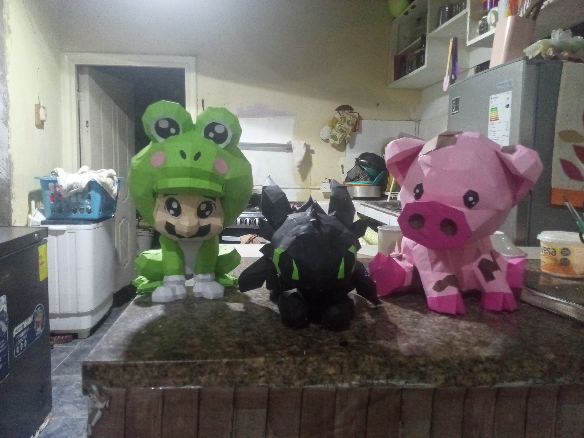 On a countertop, a polygonal figure of a person wearing a green frog suit is on the left, a black polygonal figure of a dragon is in the middle, and a pink polygonal figure of a pig is on the right. The frog and pig figures have printed faces and markings. The countertop is brown, and there is a refrigerator and other kitchen appliances in the background. The location appears to be a residential kitchen in El Valle del Espíritu Santo, Venezuela.