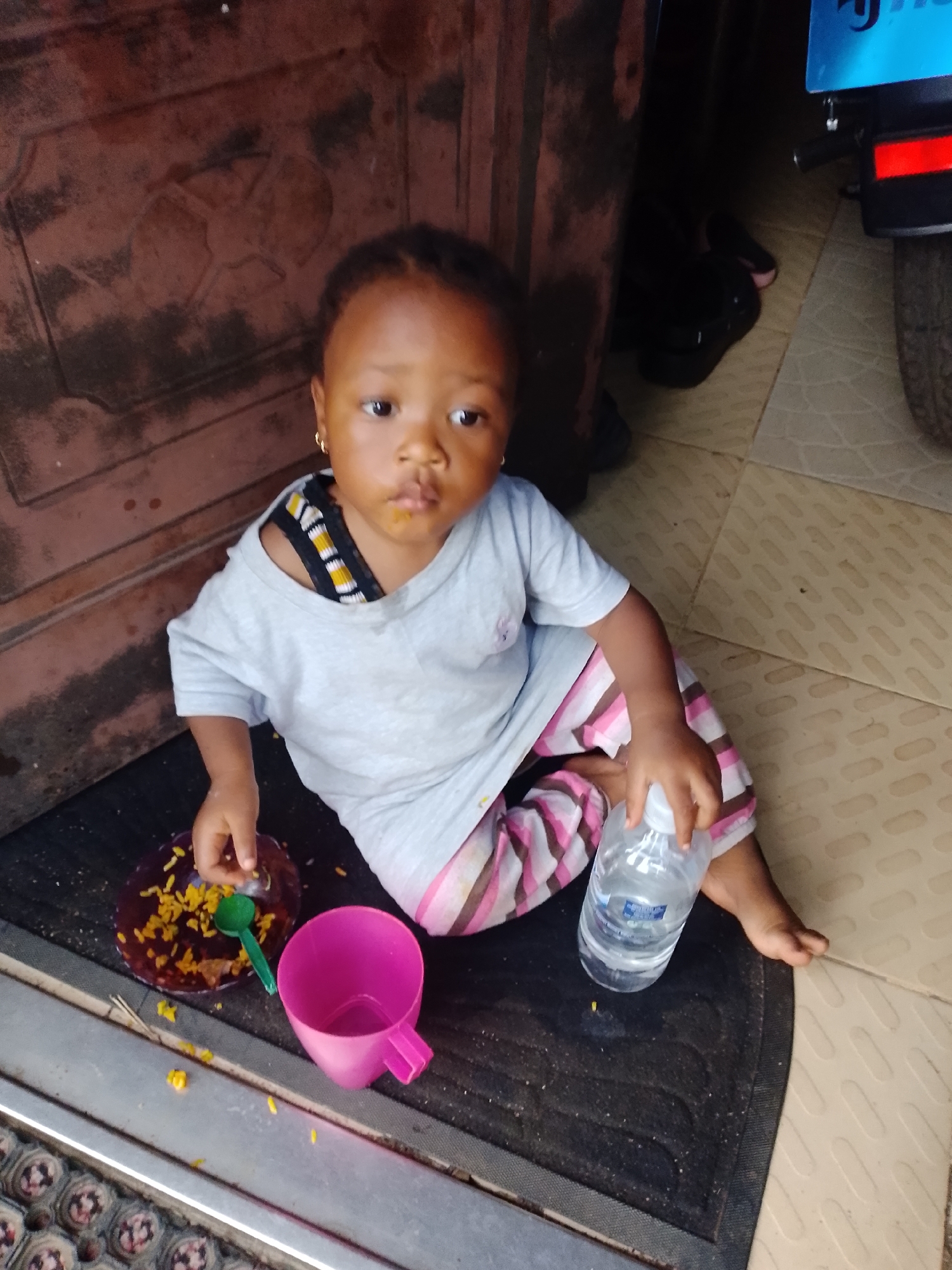 A young child sits on a black mat in front of a brown metal door. The child wears a light grey t-shirt with black, yellow, and white striped trim visible around the neckline, and striped pink and white pants. A plate of yellow rice with a green spoon sits to the child's left, and a pink cup is to the child's right. The child holds a clear plastic bottle of water in their right hand. The background shows a tiled floor and a portion of a motorcycle with a blue license plate. The scene appears to be outdoors, possibly in a residential area or near a shop in Jos, Nigeria.