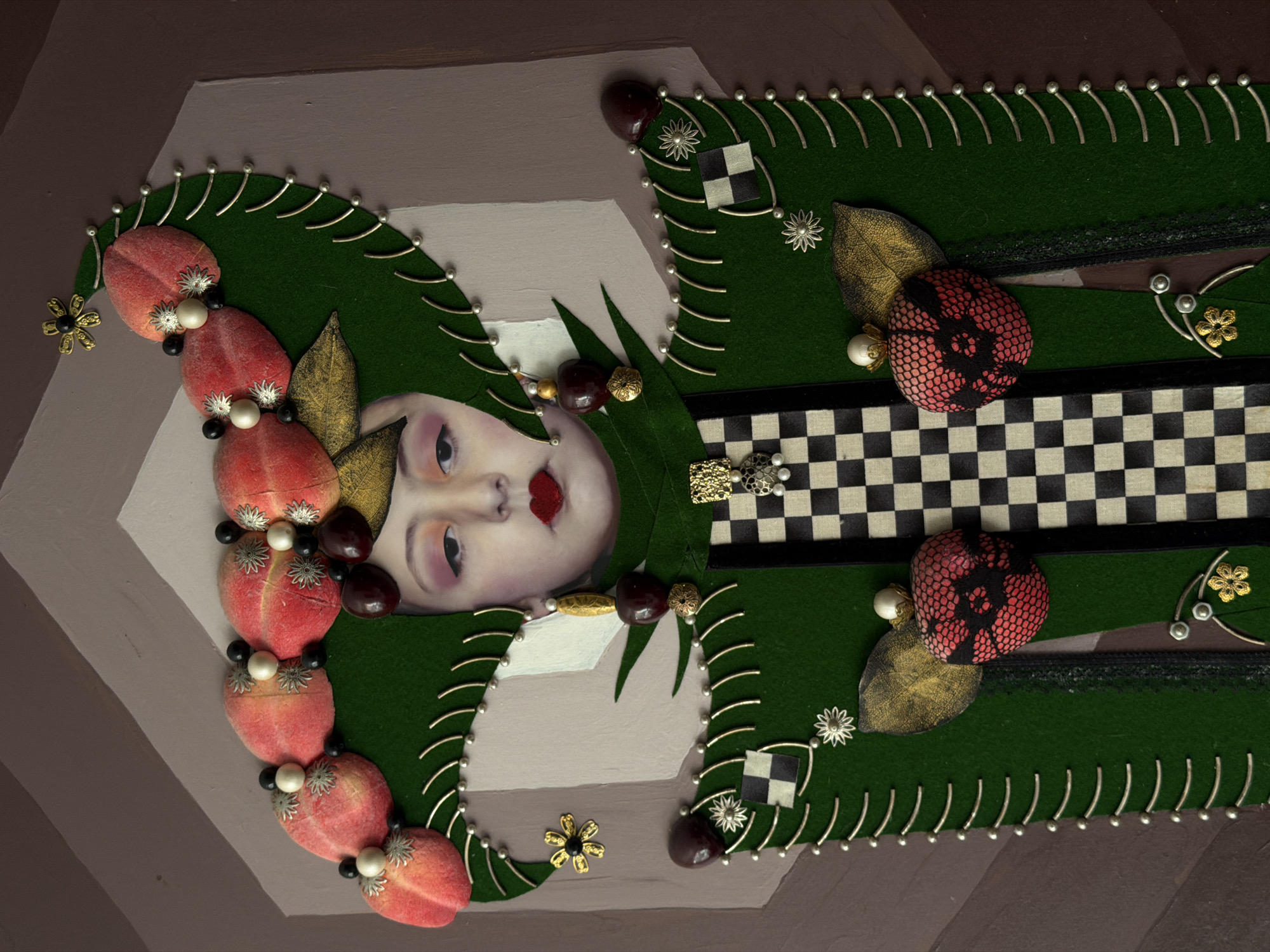 The image depicts a surreal art piece, possibly a mixed media construction or assemblage. A pale face with red lips is centrally positioned, appearing to be a collage element. It's surrounded by green felt shapes, possibly representing clothing or a headdress, decorated with silver pins, stylized floral elements, and what appear to be stylized red fruit forms embellished with beads. There are also golden leaves and checkers patterns. The background is composed of various shades of brown and grey. There is no indication as to a city or location within Yerevan, Armenia.