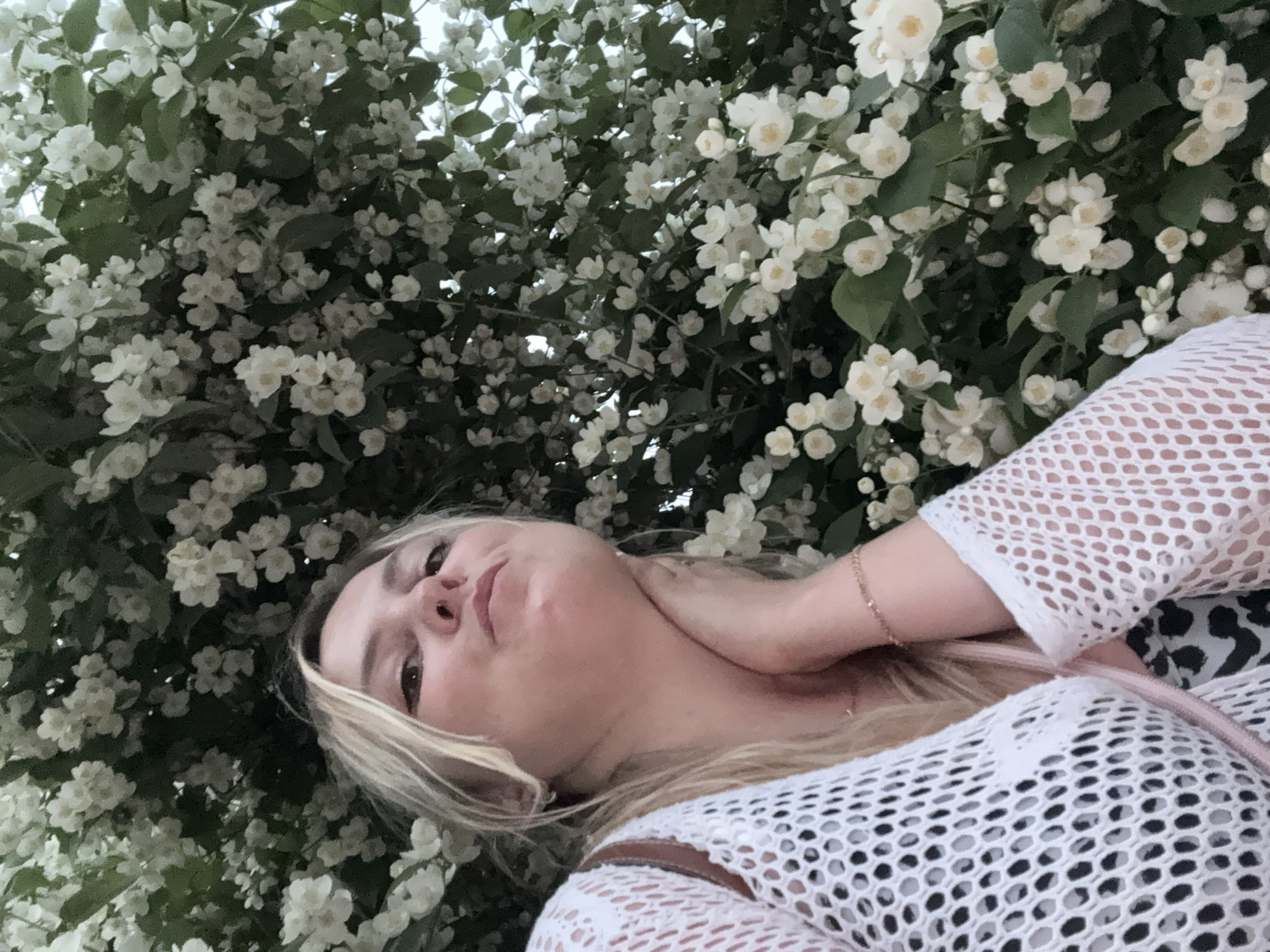 A young woman with blonde hair is taking a selfie outdoors, surrounded by white jasmine flowers and green leaves. She is wearing a white, mesh-like top over a patterned garment, and a delicate gold bracelet is visible on her wrist. The woman's face is angled upwards, with her chin slightly raised and her lips pursed. Her hand is resting on her neck. The background is filled with the dense foliage and blossoms of the jasmine plant. The image appears to have been taken during daylight, with the sky visible in the upper right corner.