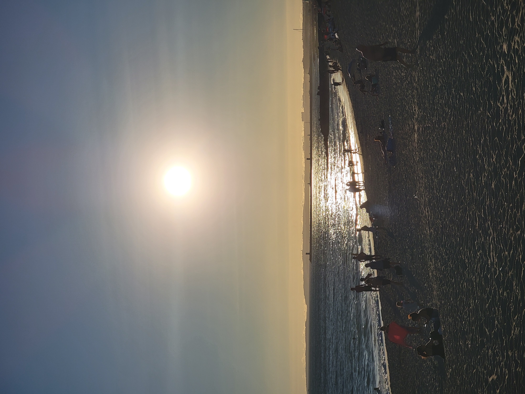 The image depicts a wide, sandy beach in Quarteira, Portugal, during the late afternoon or early evening, indicated by the low-lying sun. The sun, a bright, hazy disc, is positioned in the upper center of the frame, casting a warm, golden light across the scene and reflecting intensely on the rippling water. A distant, dark, and hazy coastline with structures and what appears to be a lighthouse or tower is visible on the horizon.
The beach is populated with numerous people engaged in various activities. Closer to the water's edge, several individuals, some appearing to be children and adults, are interacting with the sea, with some wading or standing in the shallow water. The waves are small and create a shimmering effect due to the sunlight. Further up the beach, many people are relaxing, sunbathing, or walking. There are silhouetted figures scattered across the sand, some are lying on towels, others are standing or walking. A red object, possibly a towel or flag, is visible near the water. Some people are clustered in groups, while others are solitary. The sand itself is textured with ripples and shadows, suggesting an uneven surface. The overall atmosphere is one of leisure and recreation on a summer day.
