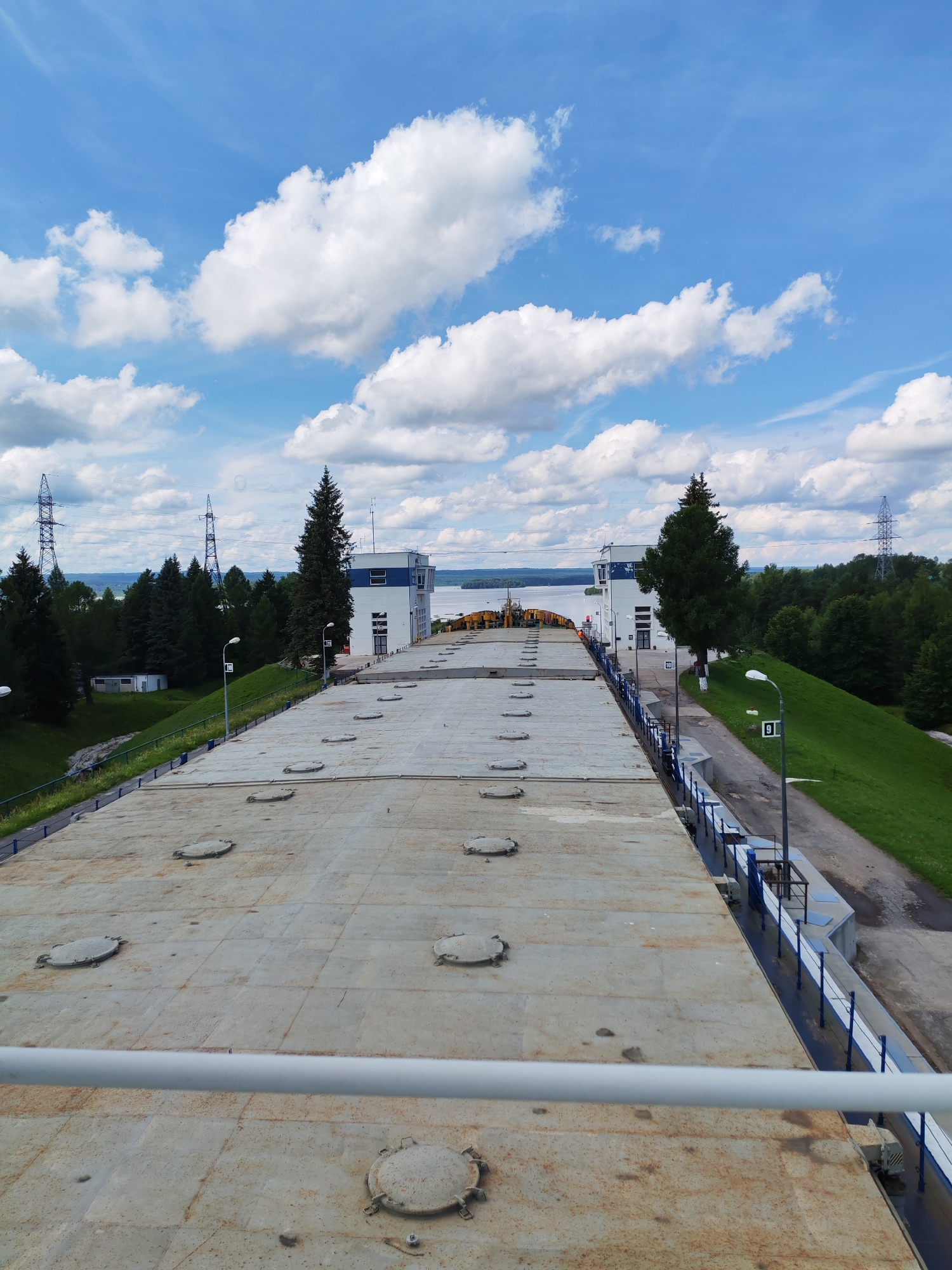 An overhead perspective shot showcases a vast, weathered, and metallic surface dotted with circular, rusty manhole covers. This expansive surface appears to be the roof of a large industrial structure, possibly a barge or a dam. A white railing runs horizontally across the foreground, framing the view of the metallic expanse. To the right of the metallic surface, a steep, grassy embankment slopes downwards, bordered by a blue and white railing. A paved walkway runs alongside the embankment, featuring a lamppost with a sign displaying the number "9".
Further in the background, two modern white buildings with blue accents stand prominently. Between them, a tall, dark green coniferous tree reaches towards the sky. Beyond the buildings and the tree-lined horizon, a wide body of water is visible, reflecting the bright blue sky dotted with fluffy white clouds. High voltage power line towers are scattered throughout the landscape, suggesting an industrial or power generation facility in the vicinity. The overall impression is one of a functional, industrial area integrated with natural elements. The location is identified as Белоусово, Russia.