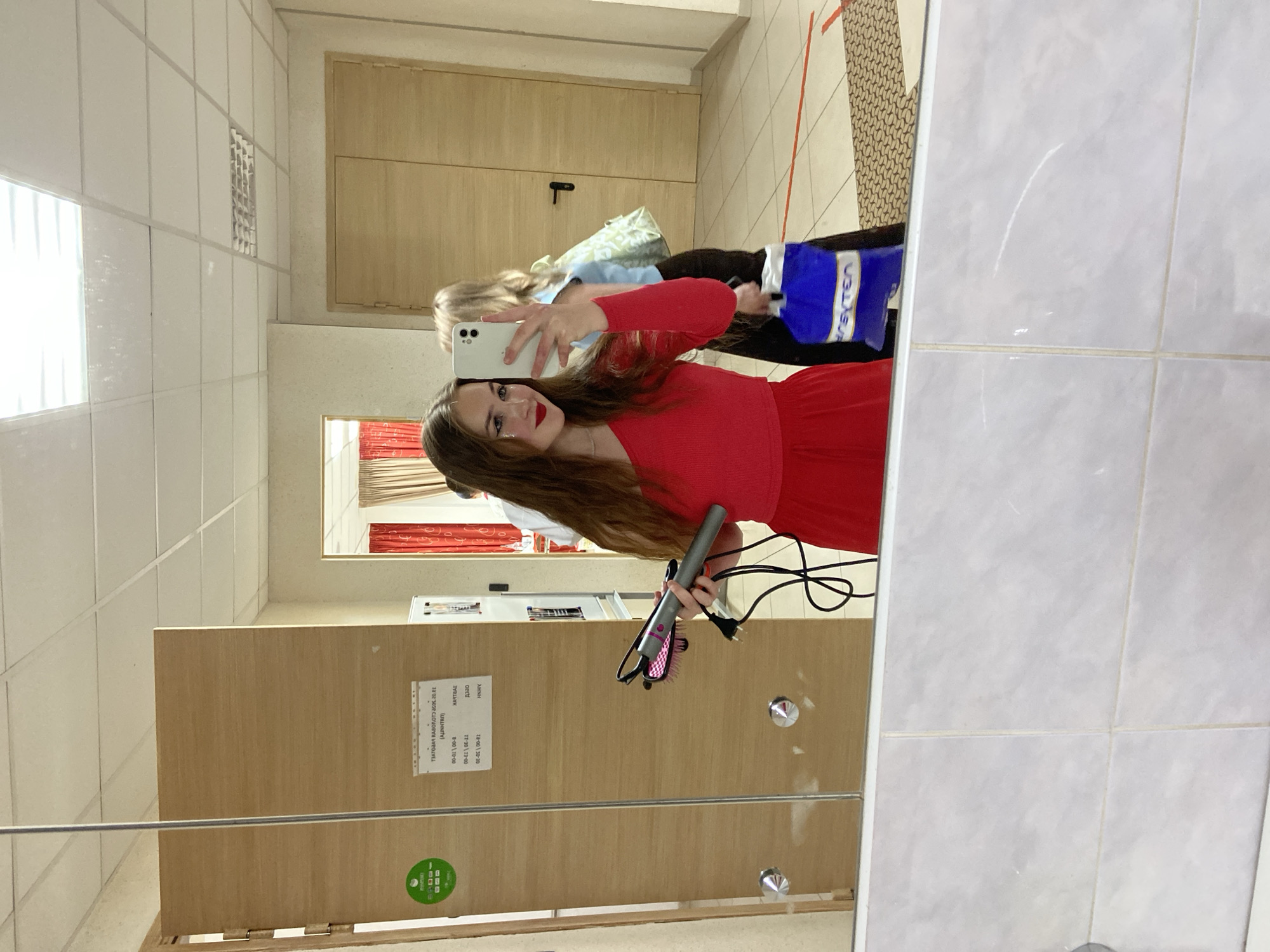 A young woman with long, brown hair and red lipstick takes a mirror selfie. She is wearing a red off-the-shoulder top and a matching red skirt. In her right hand, she holds a pink and grey hair straightener. Behind her, a blurred figure of another person is visible, wearing a light blue top and dark pants, carrying a blue shopping bag. The scene appears to be in a hallway with wooden doors and tiled walls. A sign with text is affixed to one of the doors. The location is identified as Прикамский, Russia, based on the provided context.