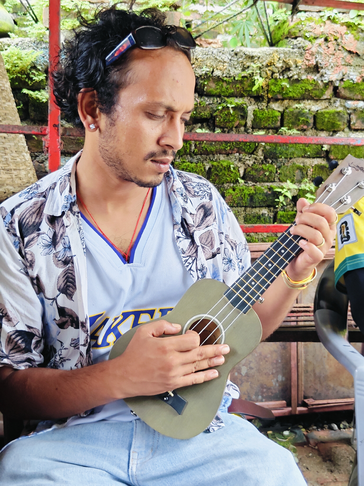 A medium shot shows a man sitting outdoors, possibly on a porch, in Pokhara, Nepal, holding a ukulele. He has dark, curly hair, a mustache, and is wearing sunglasses perched on his head and an earring. He is dressed in a floral-patterned open shirt over a blue and white v-neck jersey. The background features a moss-covered brick wall and a red metal railing, with some foliage visible. The man is looking down at the ukulele, with his fingers positioned on the fretboard. He is wearing gold rings on his right hand and a gold bracelet on his left wrist. A red string necklace is visible around his neck. The lighting suggests it is daytime.