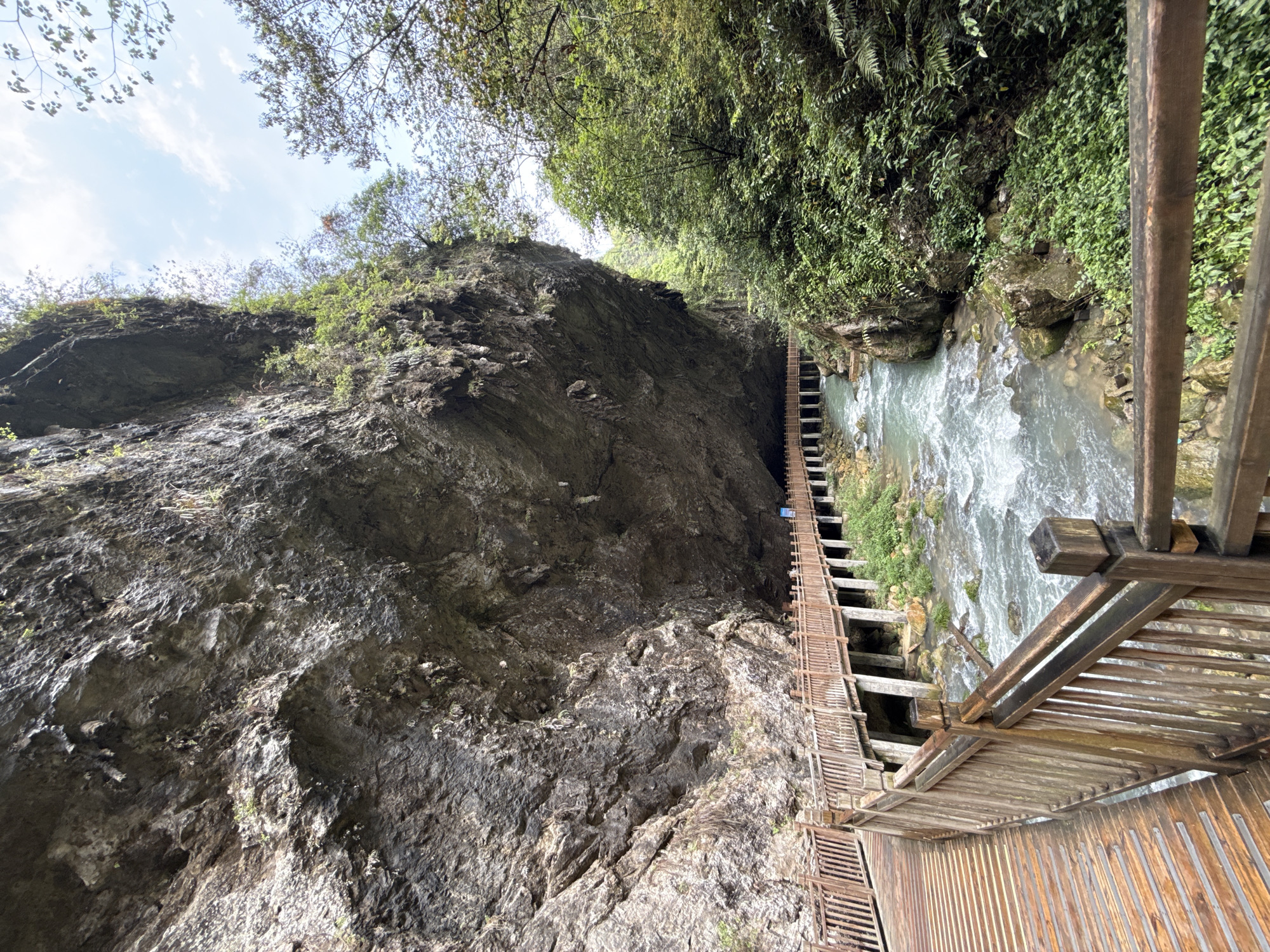 The image shows a steep gorge with a rocky cliff face on the left. On the right, dense green vegetation lines the edge of a fast-flowing river. A wooden walkway is constructed along the edge of the river, attached to the cliff face in places, with wooden railings visible in the foreground. This walkway is located in the city of Zhangjiajie, China.