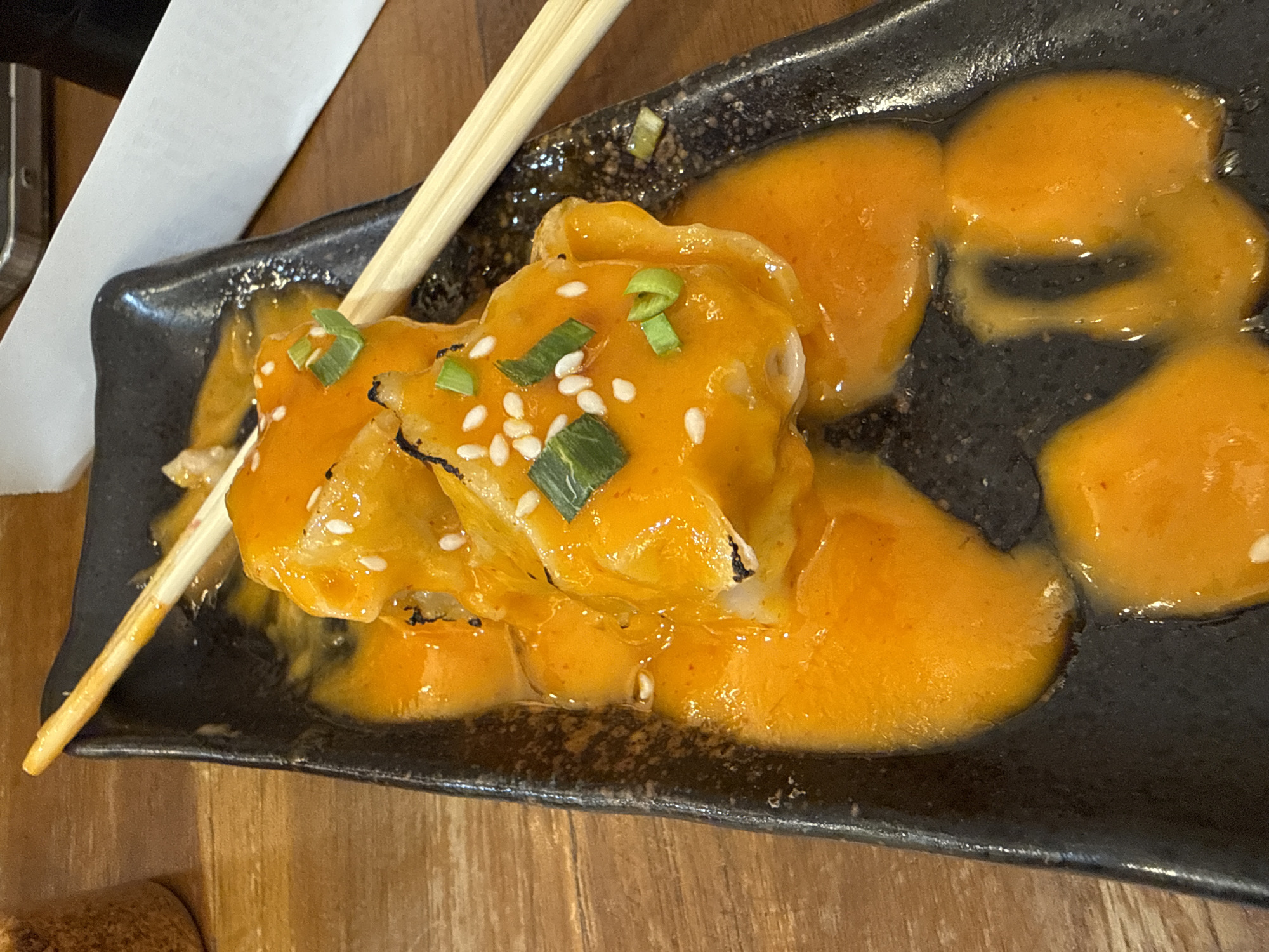 A plate of dumplings sits on a wooden surface, potentially a table. The dumplings are covered in an orange sauce and garnished with sesame seeds and chopped green onions. A pair of wooden chopsticks rests on the plate. The plate itself is black and has a rough, possibly textured surface. The context suggests the image may have been taken in Lhokseumawe, Indonesia, although this is not evident from the image alone. The focus is on the presentation of the food. No people are visible.