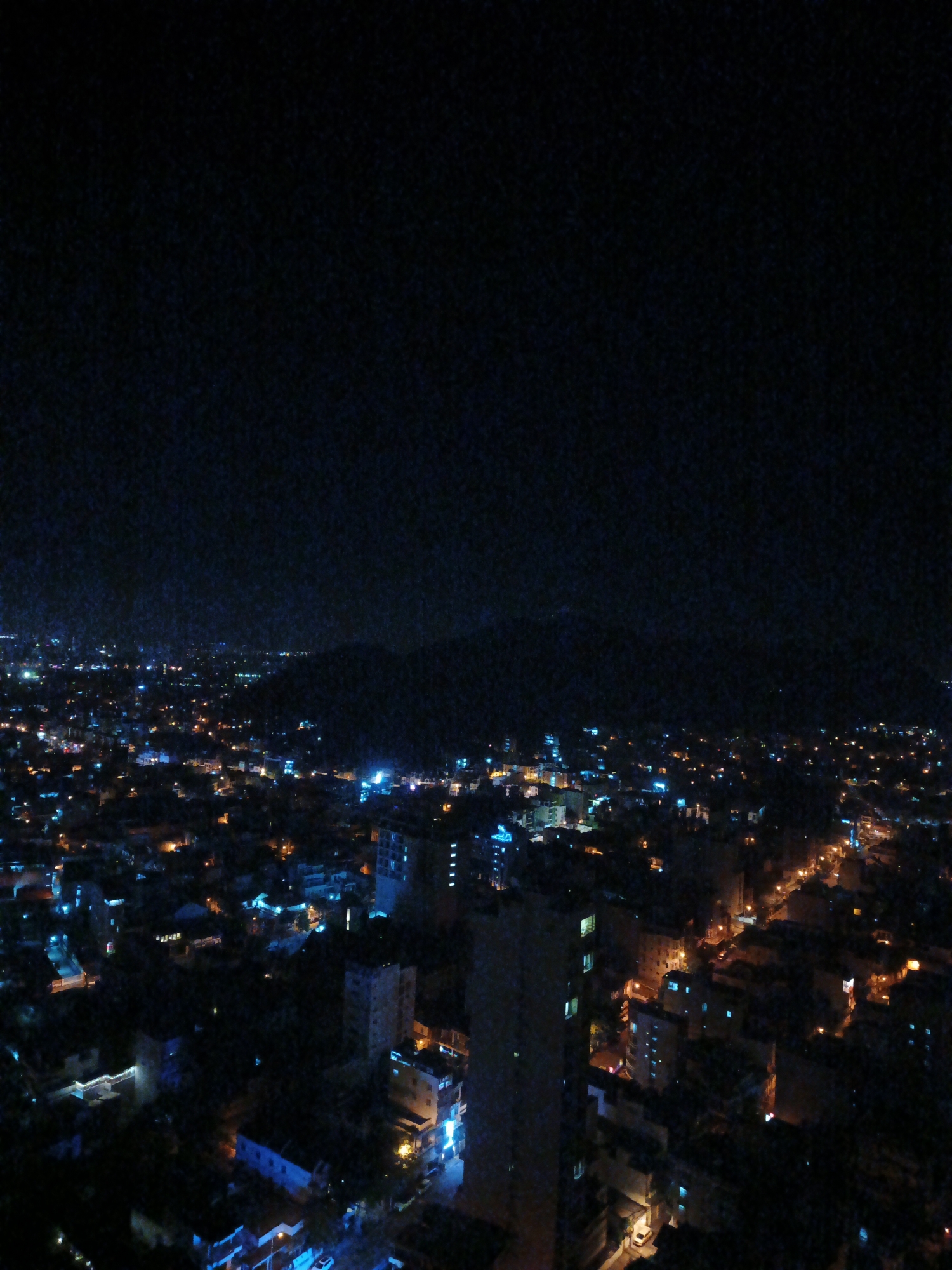 The image depicts a nighttime aerial view of a dense urban area, likely Bac Nha Trang Ward in Vietnam, characterized by a sprawl of buildings illuminated by artificial lights. The sky is dark and appears to be filled with a subtle grain or noise, obscuring any celestial bodies. Below, the cityscape is a mosaic of lights of varying colors, including bright white, blue, and orange, emanating from windows, streetlights, and building facades. The terrain appears to be relatively flat in the foreground, with a darker, elevated area that could be a hill or mountain range visible in the distance. The scene is devoid of any visible people or vehicles, focusing solely on the urban infrastructure and its illumination against the night. The buildings are a mix of low-rise and multi-story structures, some appearing to be apartment complexes or commercial buildings. The overall impression is one of a bustling city at rest, with its lights painting a pattern across the darkened landscape.